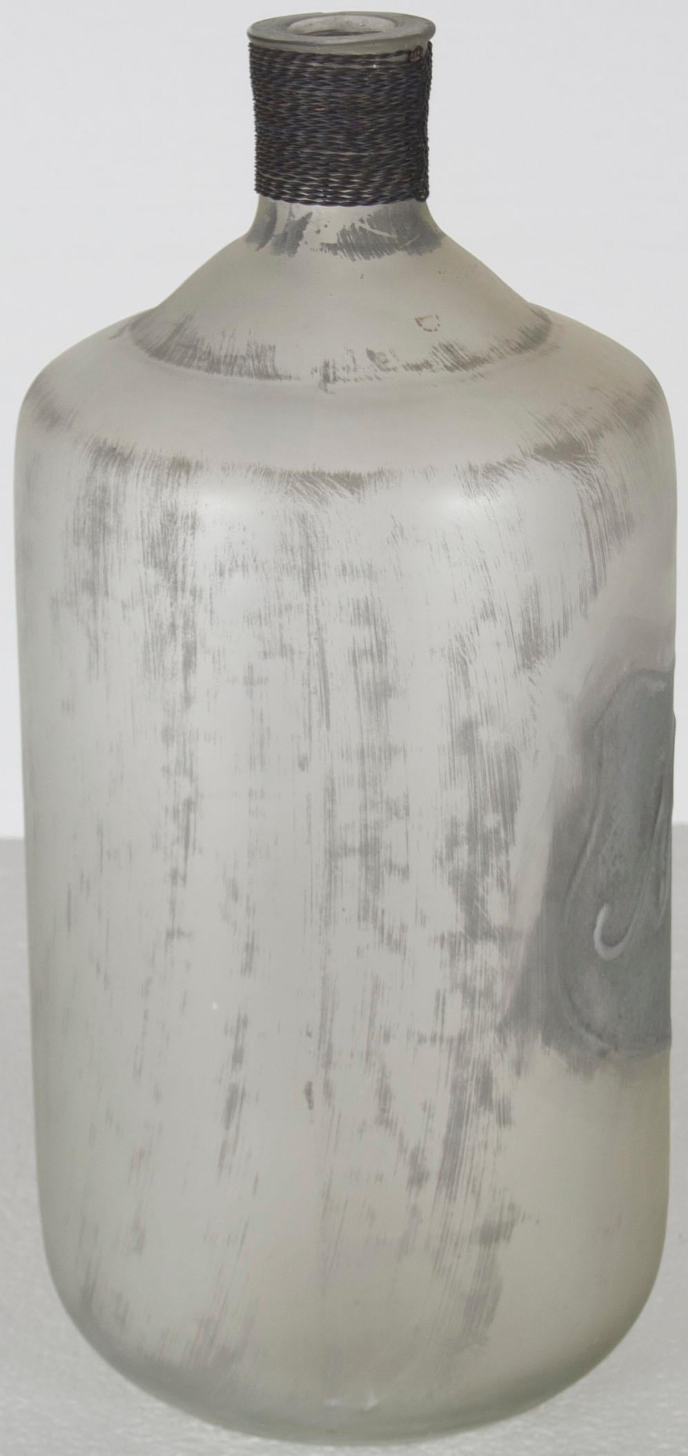 This large frosted glass jug is a new item, made like wine jugs of old. The glass is a distressed, frosted white with grey. The neck has been wrapped in a metal wire cord for a subtle contrast. A shield emblem with “No 3? has been embossed on the
