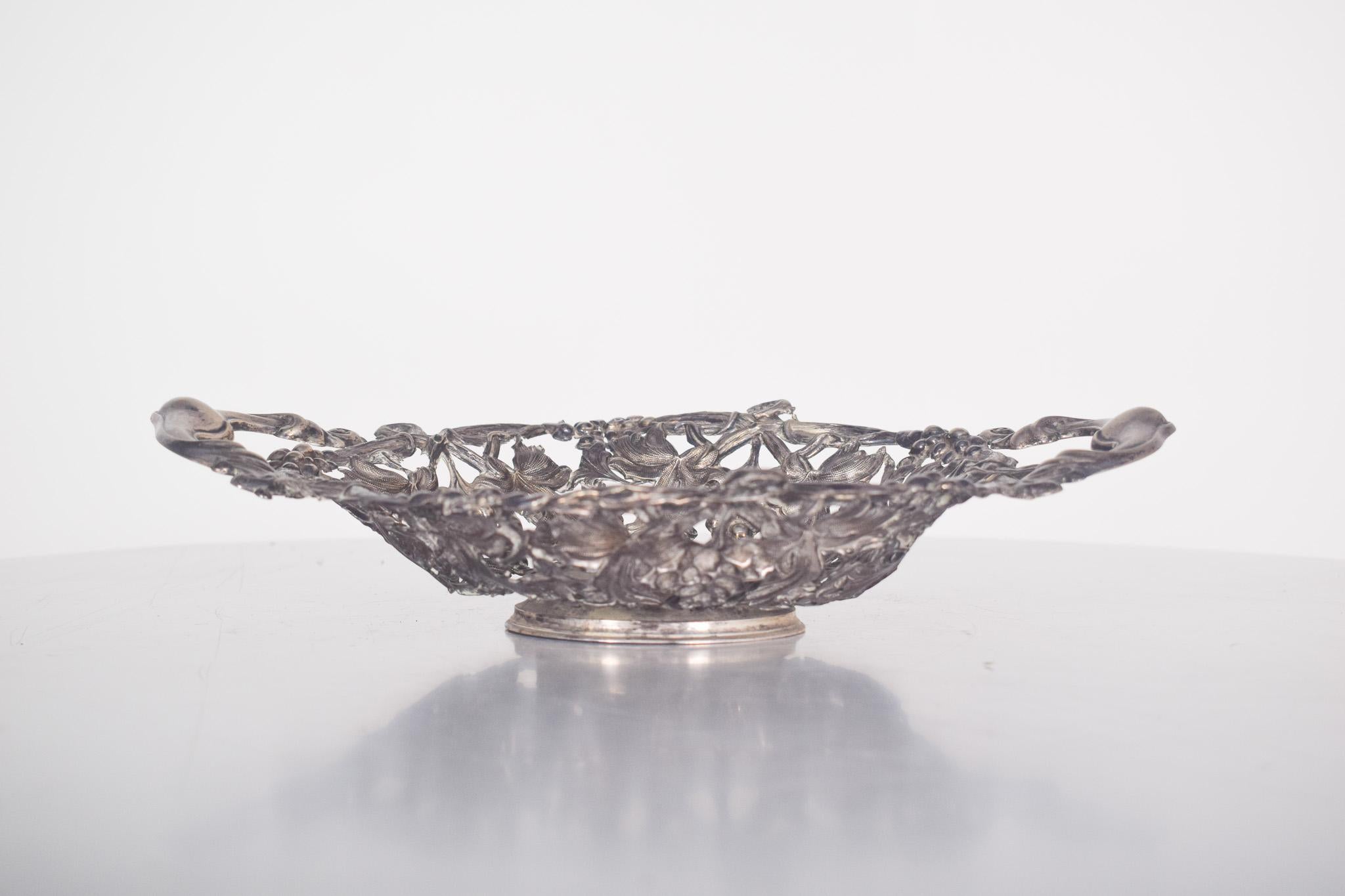 Obstschale aus durchbrochenem und bearbeitetem Silber, mit zwei Henkeln. Reich verziert mit Reliefs, Weintrauben und Weinblättern. 
Mit zwei eleganten, geschwungenen Griffen zeigt das Stück ein raffiniertes und funktionelles Design. Auffallend ist