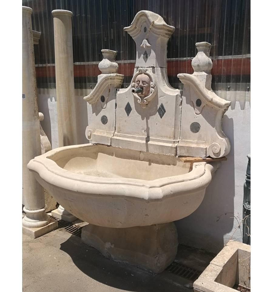 Italian wall fountain of the 18th century made of stone, with marble incristations making shapes of rhombuses and circles, with a marble mask of reddish verona in the central part, fountain of classic style.