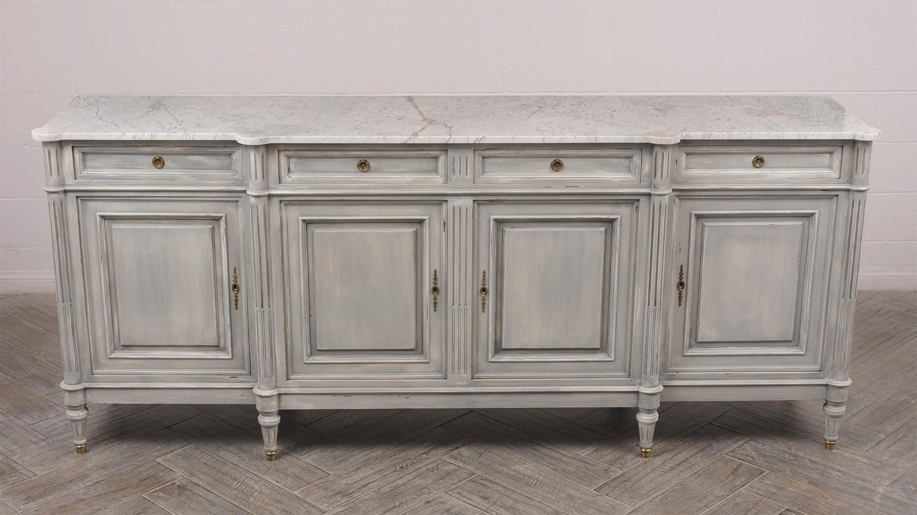 This 1950s French Louis XVI style buffed is made of solid cherrywood that has been painted in a pale grey color with a distressed finish. The buffet features a white marble top w/gray veins. Four side-by-side top drawers with center circular brass