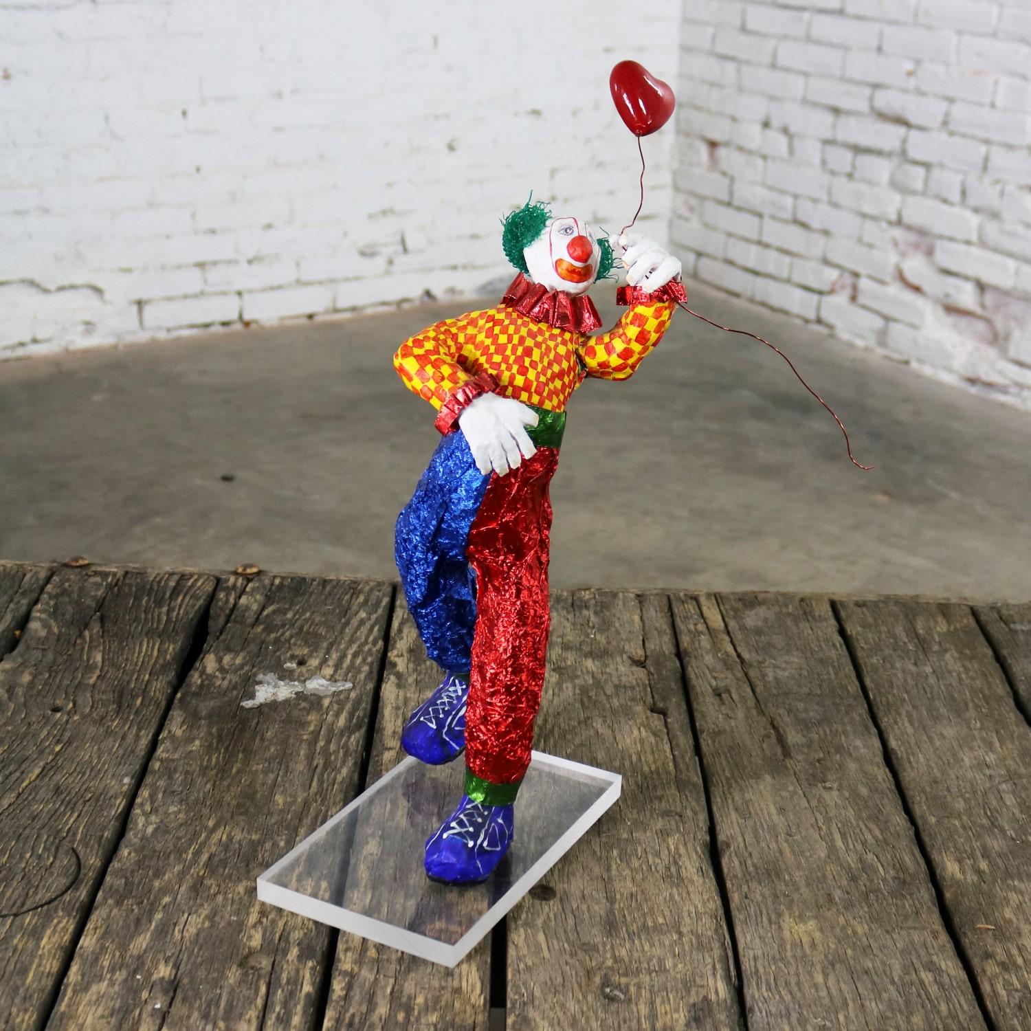 Fun and funky and bright Folk Art clown sculpture carrying a heart balloon of mixed-media including paper maché, paper, plastic, and wire on a plexiglass stand. It is in wonderful vintage condition. There is a repaired crack on one of his underarms,