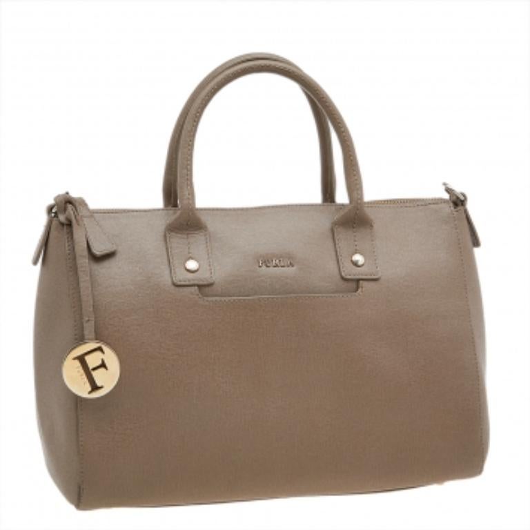 Este elegante bolso tote Linda es de la casa Furla. Fabricado en piel y forrado de tela en el interior, el bolso se completa con dos asas superiores y un colgante con el logotipo. Llévala de compras o al trabajo.