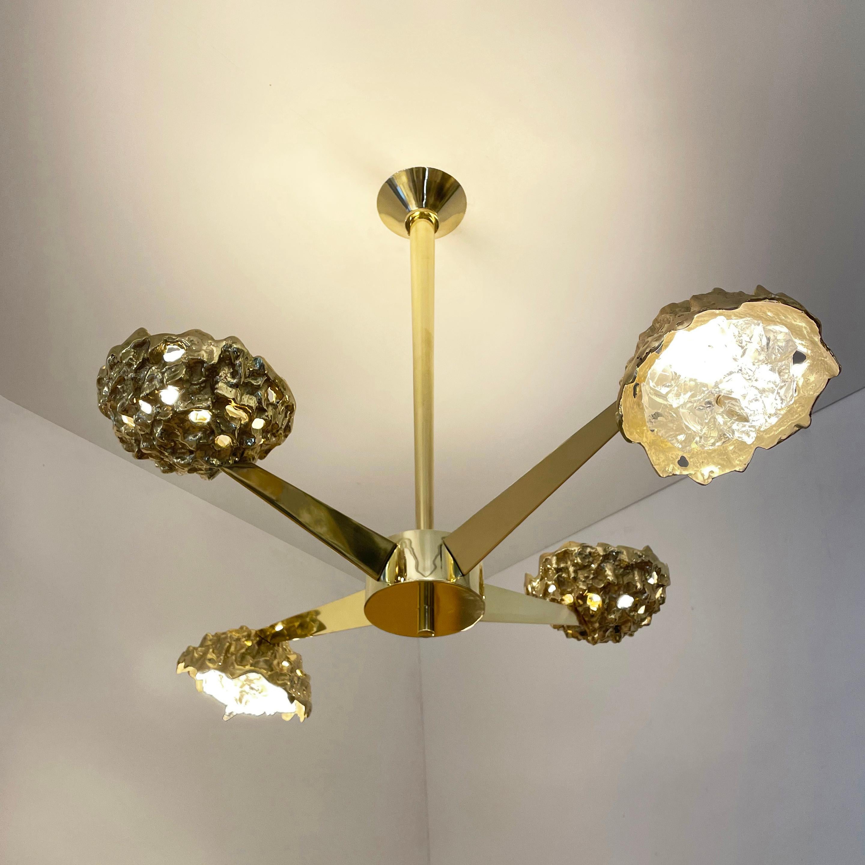 La plafoniera Fusione N.4 mette in risalto il contrasto tra forme organiche e linee geometriche eleganti; i paralumi in ottone fuso con cristalli di vetro irregolari spuntano da bracci a forma di V lucidati. In ottone lucido e vetro trasparente.