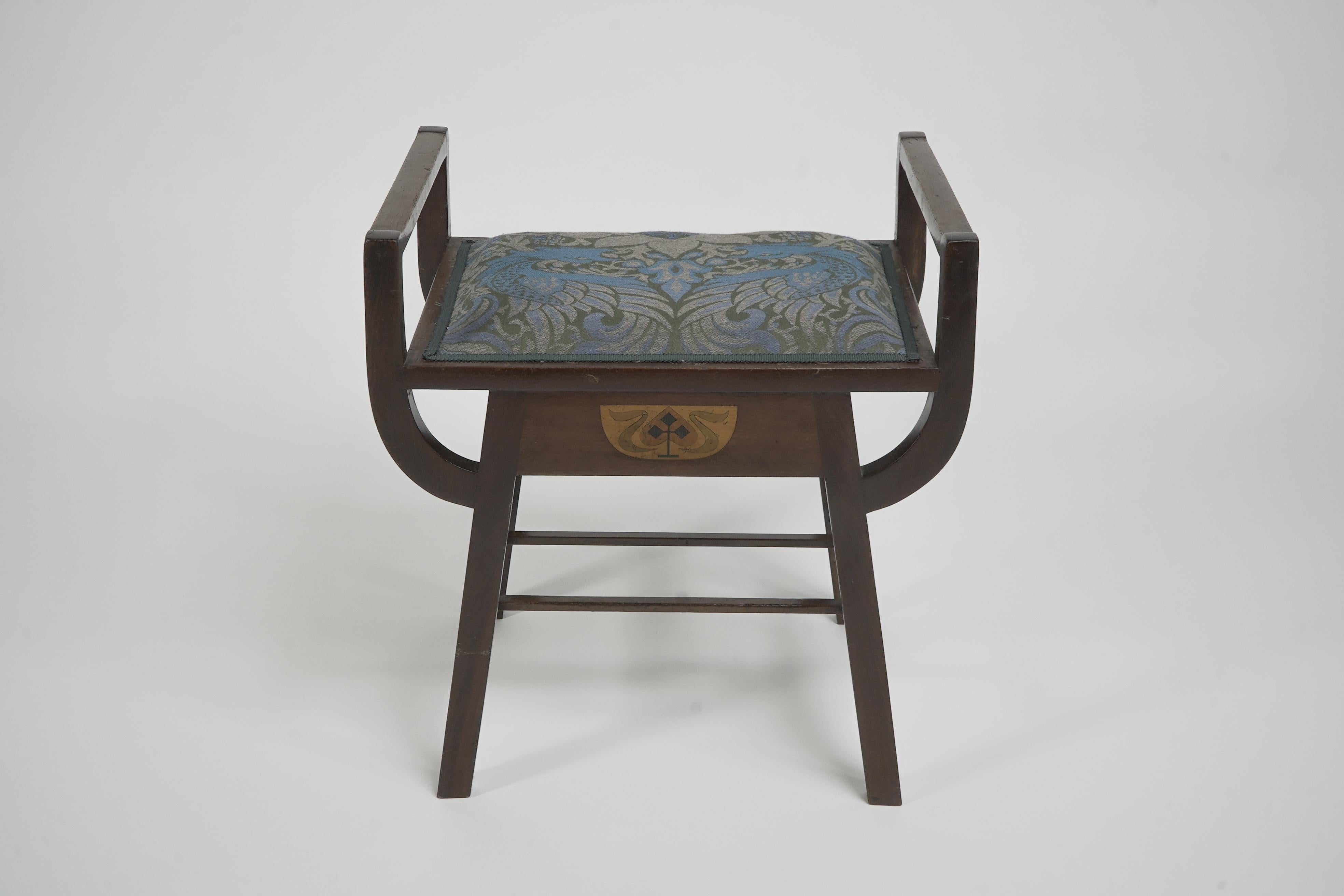 G M Ellwood and made by J S Henry. 
An Arts and Crafts 'A' framed piano or dressing table stool, with raised sides that simultaneously unite with the legs and the seat sides, a nice progressive design detail. Inlaid with a stylised motif to the