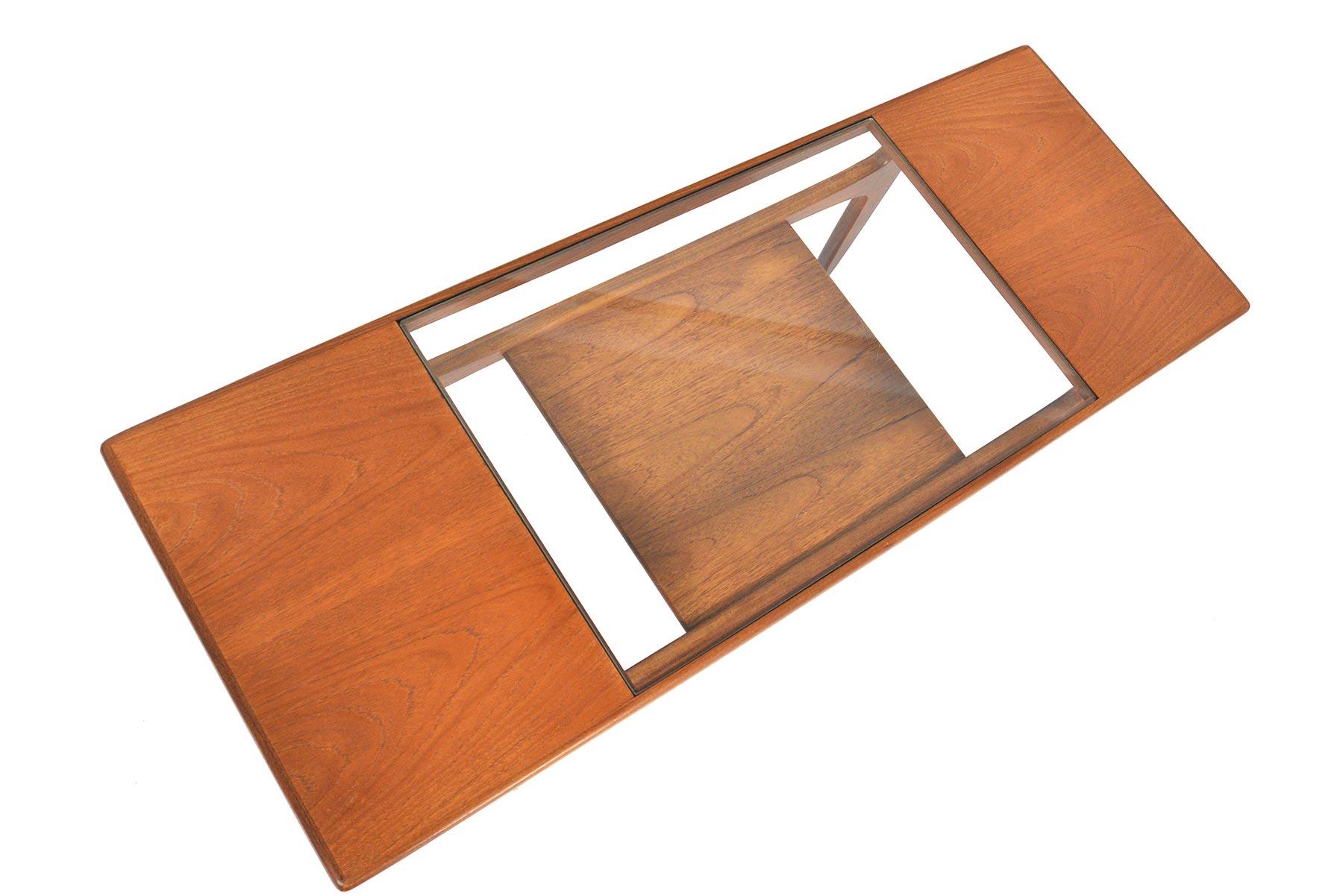 This English Mid-Century Modern teak and glass surfboard coffee table was designed by Victor Wilkins for G Plan’s Fresco range. The glass insert provides coaster free entertaining, and features a lower teak rack for books and magazines. In excellent