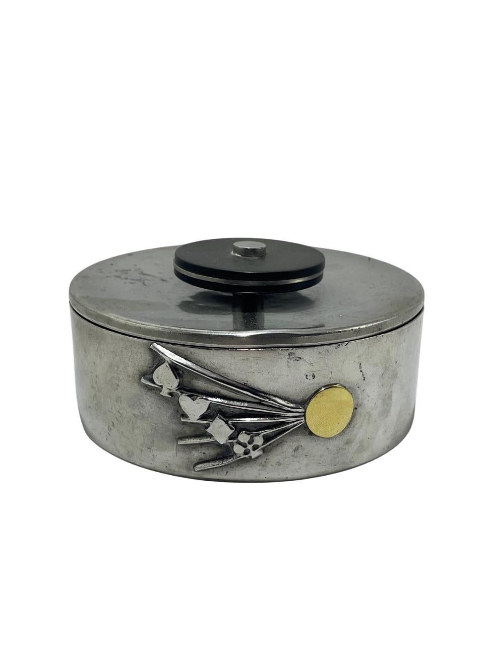 Esta caja vintage de peltre GAB, procedente de Suecia, presenta un diseño único, con un estilizado estallido de palos de naipes -pada, corazón, diamante y trébol- que irradian desde un acento circular dorado. La elegante caja de forma ovalada tiene