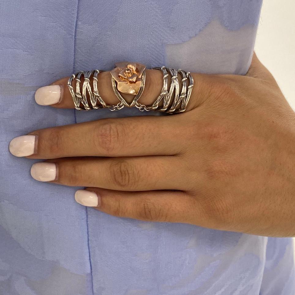 A 10k rose gold rosebud blooming atop the knuckle connects two separate sterling silver rings created of multiple vines that crisscross over one another. This iconic design is sculpted in mixed metals to span the length of the finger adjusting