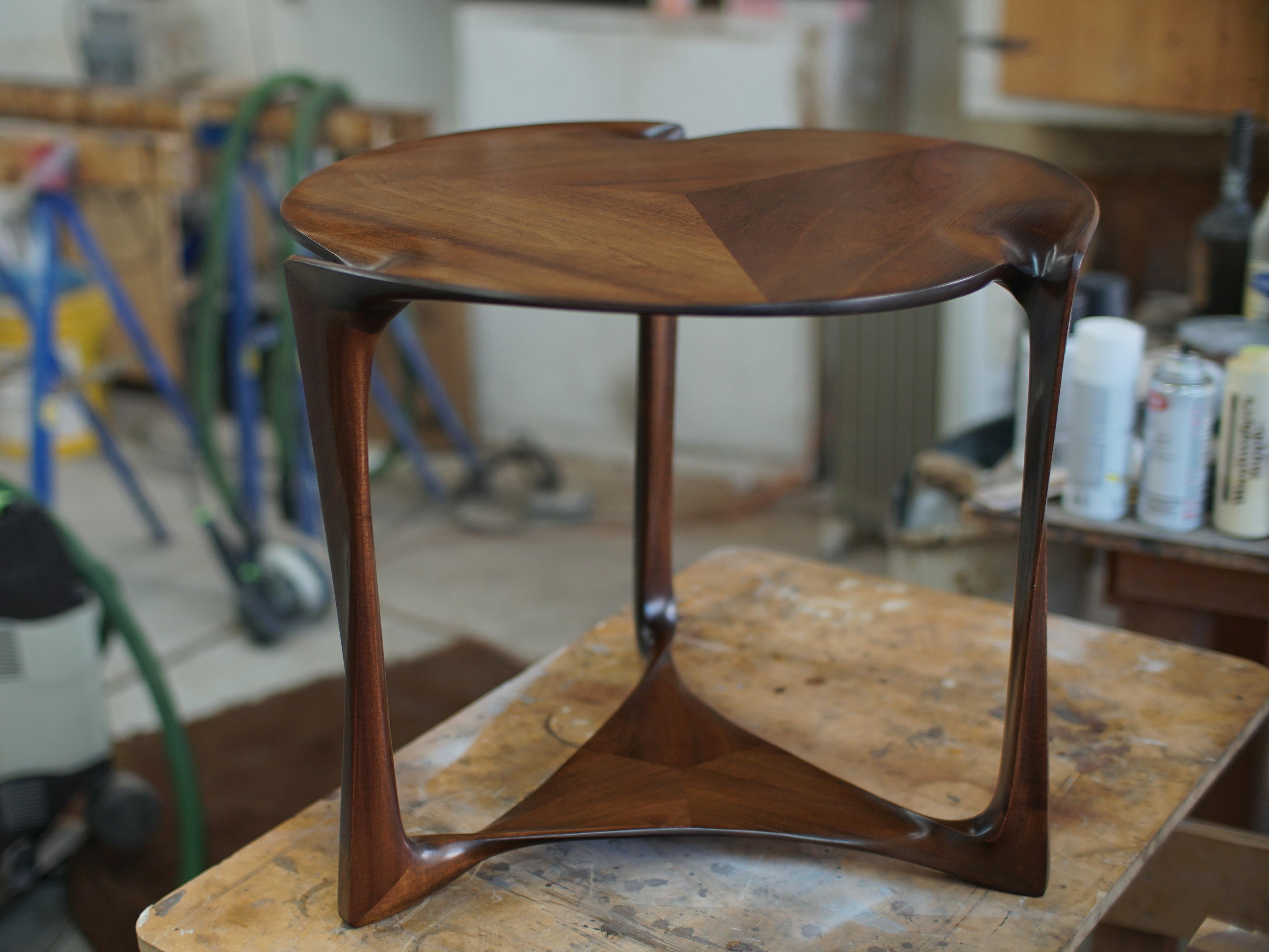 The Classic geometry of this little table, a round top over a triangular shelf or stretcher assembly, supported by three legs, is given a sculpted, organic form employing traditional carving techniques not seen since the great masters of Art