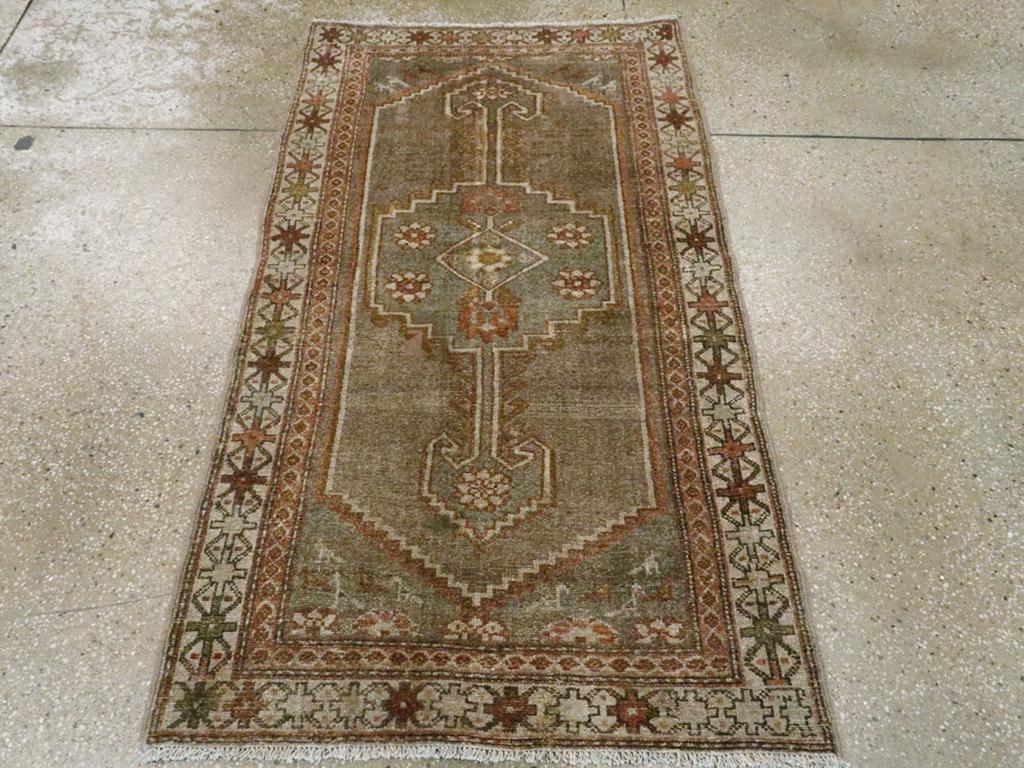 Esta alfombra persa Malayer de principios del siglo XX, que mide 1,80 m x 1,80 m, presenta un diseño audaz y simétrico centrado en torno a un medallón alargado con bordes escalonados y rosetas florales. El campo de color oliva apagado sirve de sutil