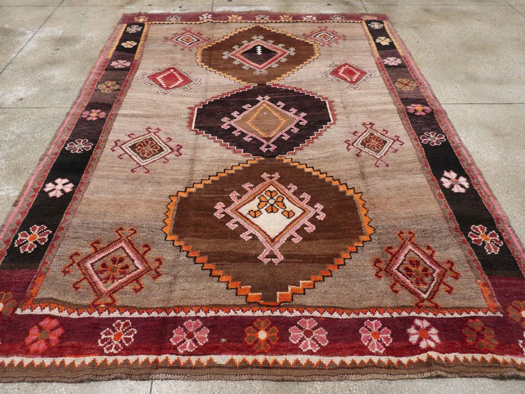 Esta encantadora alfombra tribal turca de Anatolia de mediados del siglo XX, de tamaño pequeño para una habitación, mide 2,5 m x 2,8 m y presenta una llamativa disposición de tres grandes rombos en forma de gancho que atraviesan verticalmente el