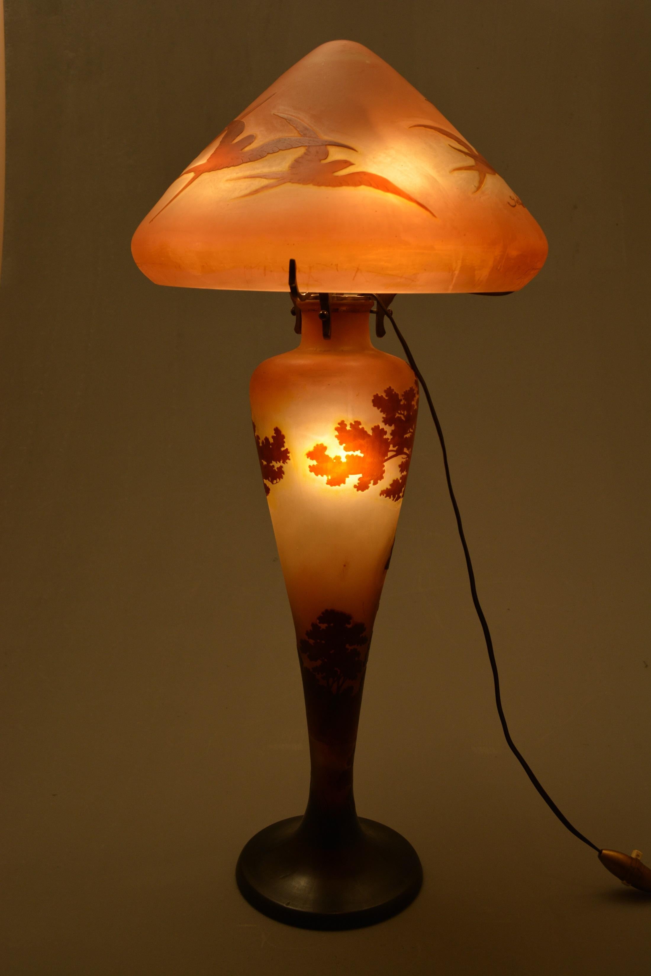 Double-overlay glass table lamp, the conical shade of translucent salmon coloured glass overlaid in a pale brown is etched with swallows with cameo signature,
The trumpet shake base of the same glass etched with a Lorraine landscape with cameo