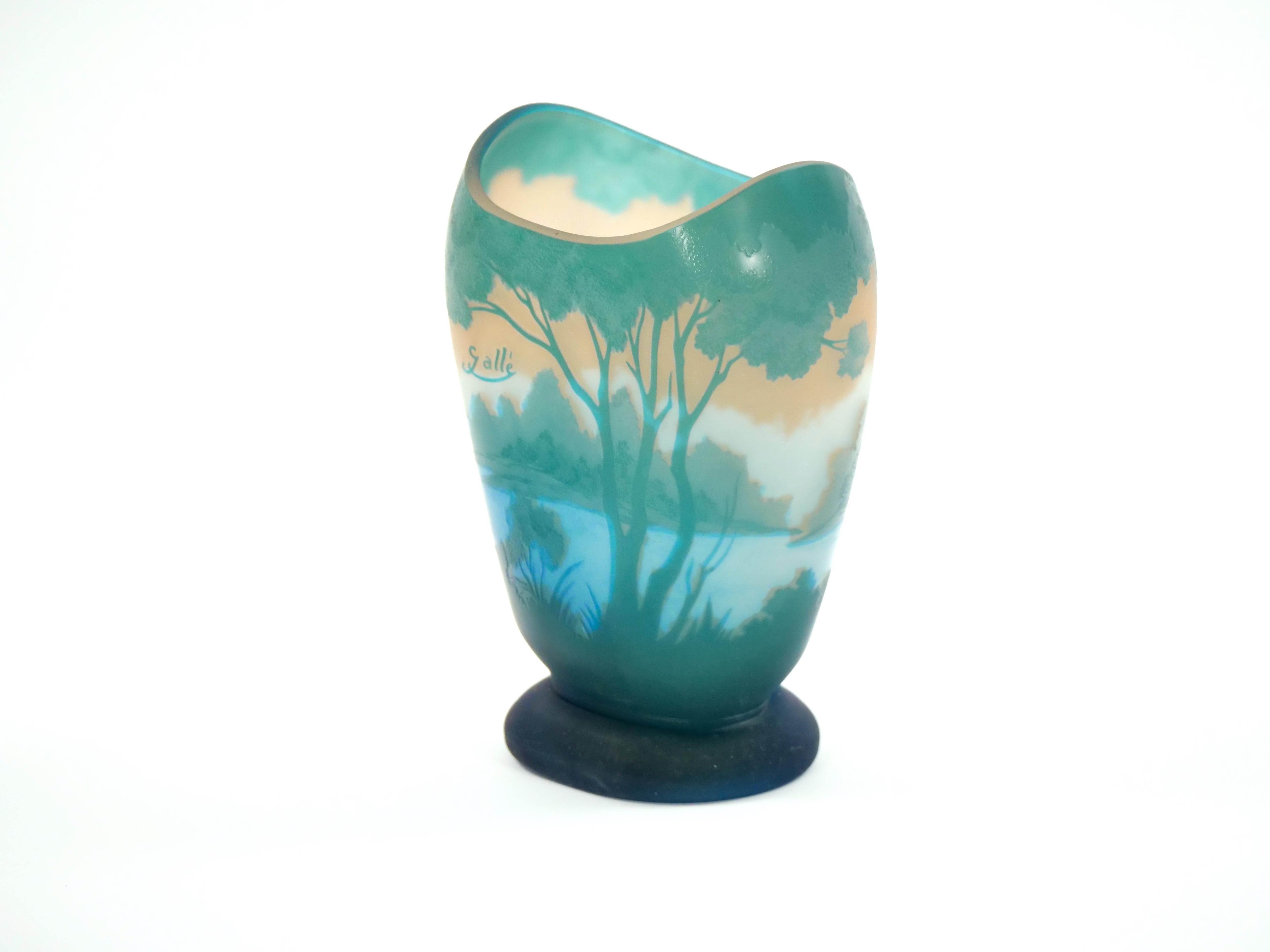 experience the beauty of nature and artistry with this exquisite Galle cameo glass vase. This masterpiece captures the serene charm of a lakeside wooded landscape, rendered in a harmonious palette of tranquil turquoise, warm tan,  and gentle light