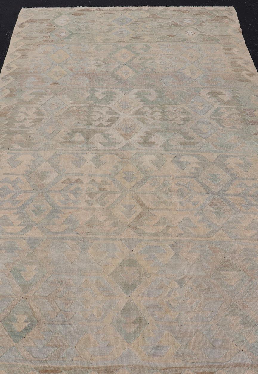 This vintage Turkish flat weave Kilim features tribal relics and faded earthy neutrals. The repeating design covers the entire field of muted light blue, caramel, taupe and gray. The border is a tribal saw-tooth design wrapping around the entire
