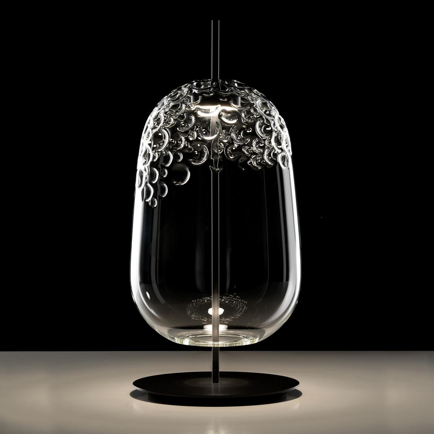 Suspension and table lamp in clear blown glass with opaque anthracite painted metal frame. The precious handwork made it possible to obtain an irregular texture that recalls the manufacturing of shark tanned leather, also called 