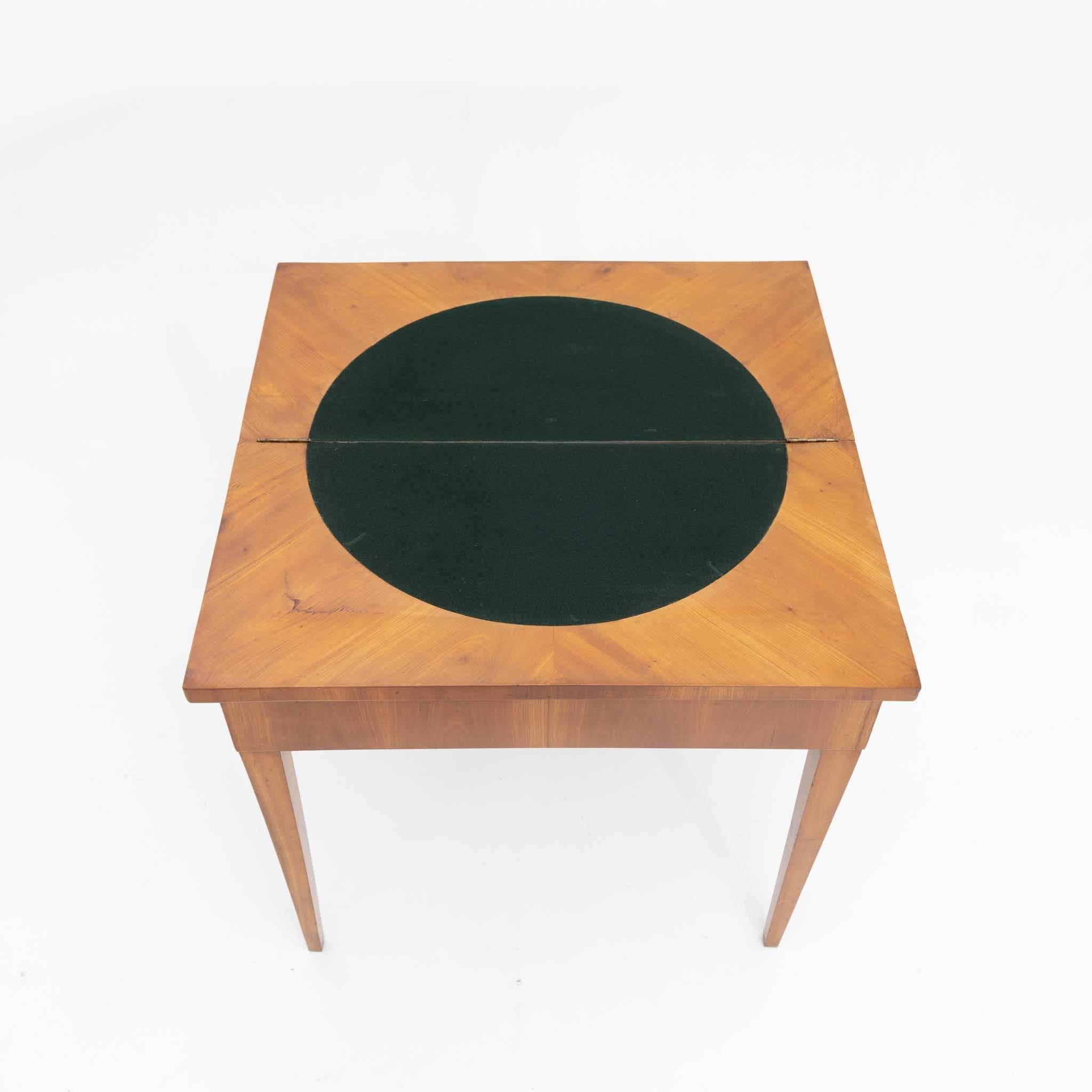 Cherry veneered game table with fold-out square tabletop with circular felt cover. The back legs can be pulled out so that the unfolded tabletop rests stably. There is storage space underneath for anything a game night needs.