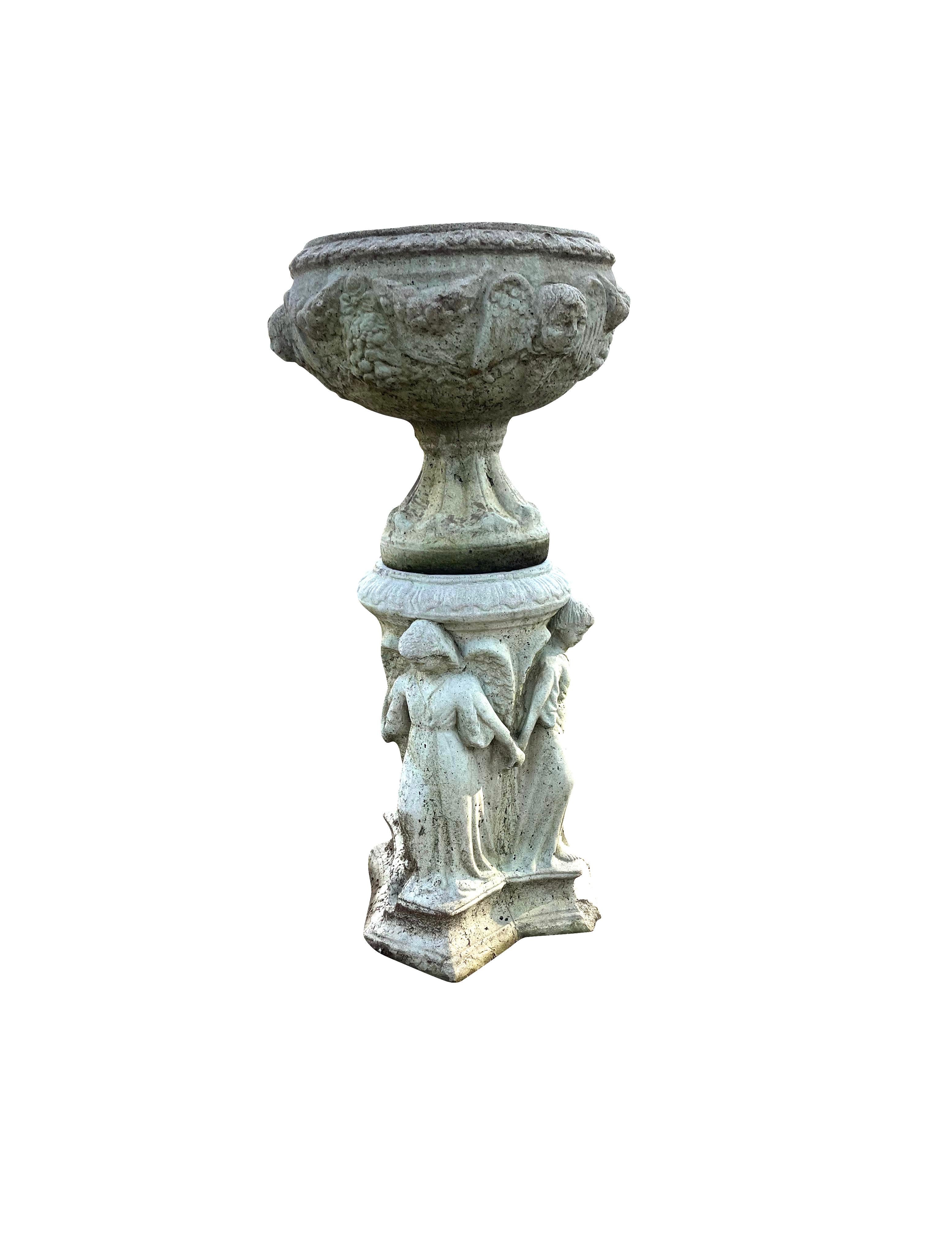 This font/ urn on a pedestal is two piece garden element consisting of a cement font on top decorated with angel or putti faces and garland on a footed pedestal base. The font/ birdbath/ planter sits on a cement base surrounded by figures of angels