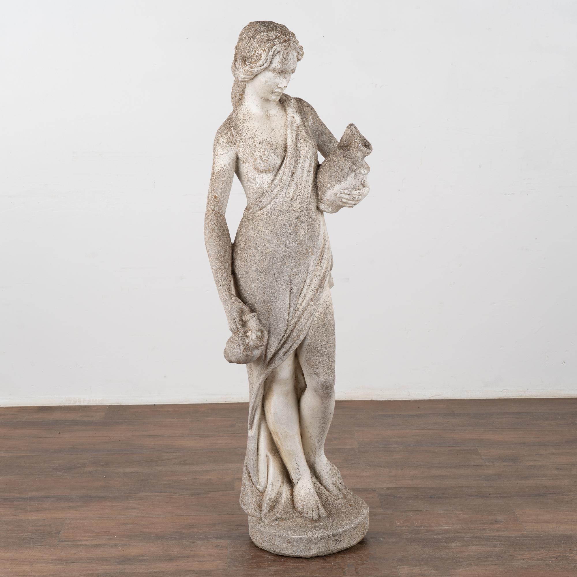 Carved limestone garden sculpture, figure of standing female with wine jugs on capital. 
The worn patina reflects years of exposure outside in the elements, leaving an aged pitted patina with lichen/stains throughout as well. 
Provenance: A Danish