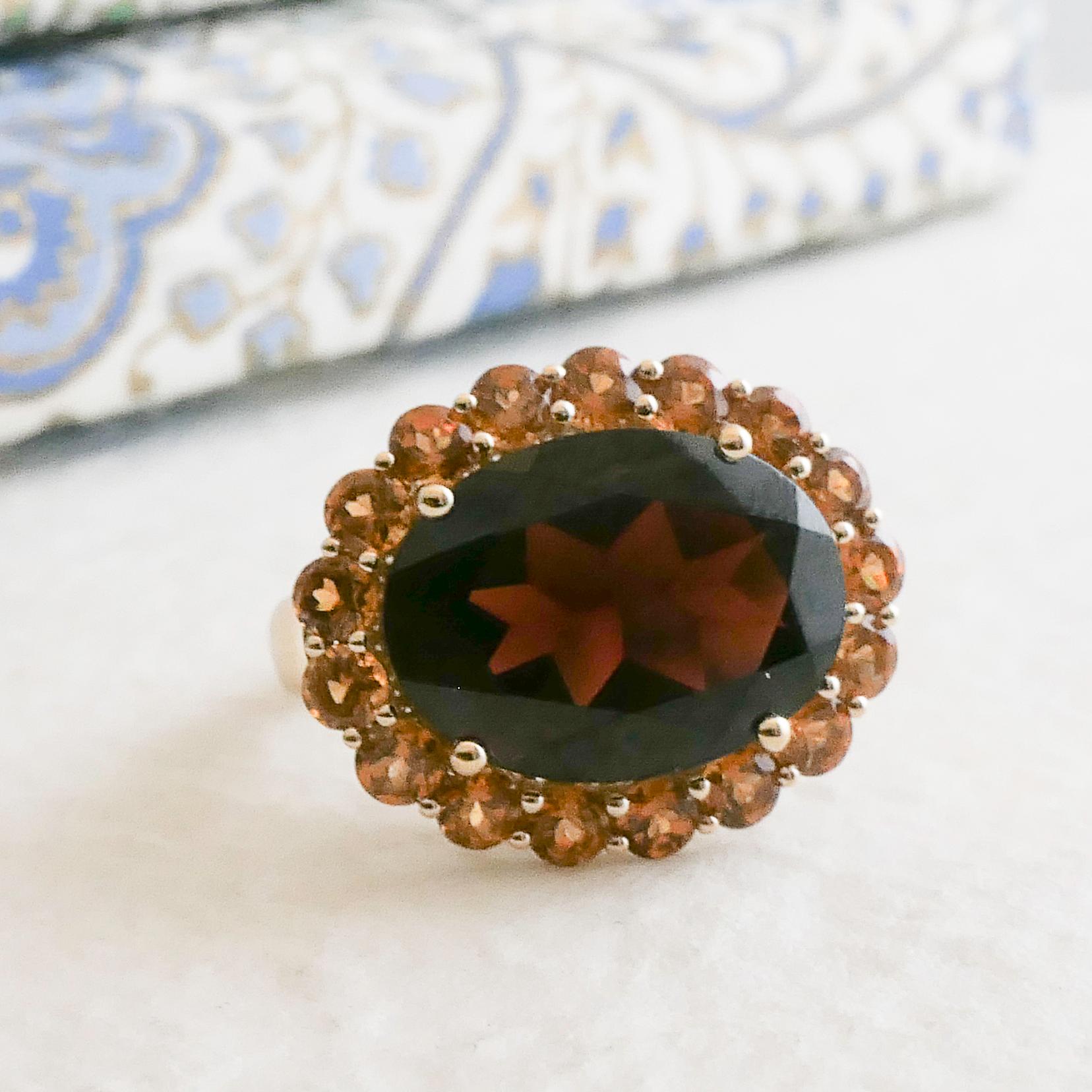 Cette bague étonnante présente en son centre un magnifique grenat ovale de 12x16 mm, aux teintes rouges profondes et à l'éclat captivant. Le grenat est entouré d'un halo de pierres précieuses en hessonite de 3 mm, ajoutant des accents chauds de brun