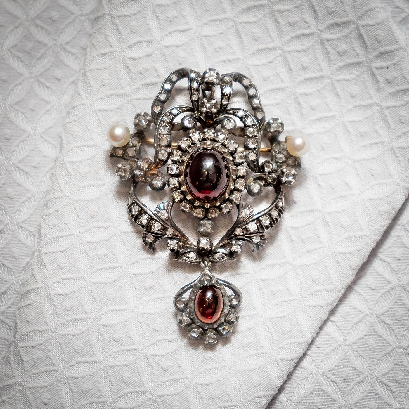 A garnet, pearl and rose-cut diamond brooch-cum-pendant, with a centrally set cabochon garnet, with a rose-cut diamond set cluster, in an openwork diamond set bow and foliate surround, with a natural pearl either side and a garnet and rose-cut