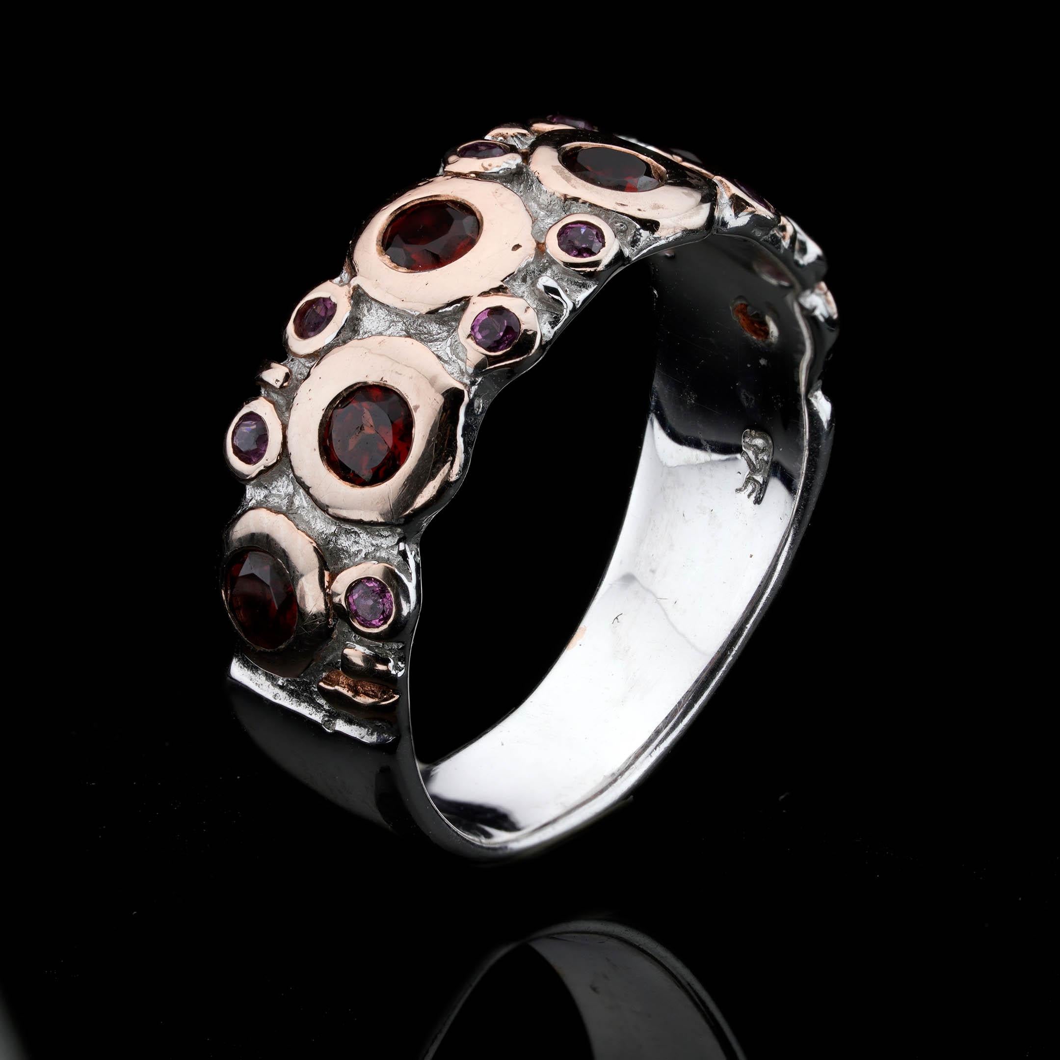 Cette bague audacieuse, fabriquée de manière artisanale, présente une bande dynamique de grenats d'un rouge profond et de saphirs roses étincelants, sertis dans une base d'argent sterling fortement texturée avec de riches accents de superposition en