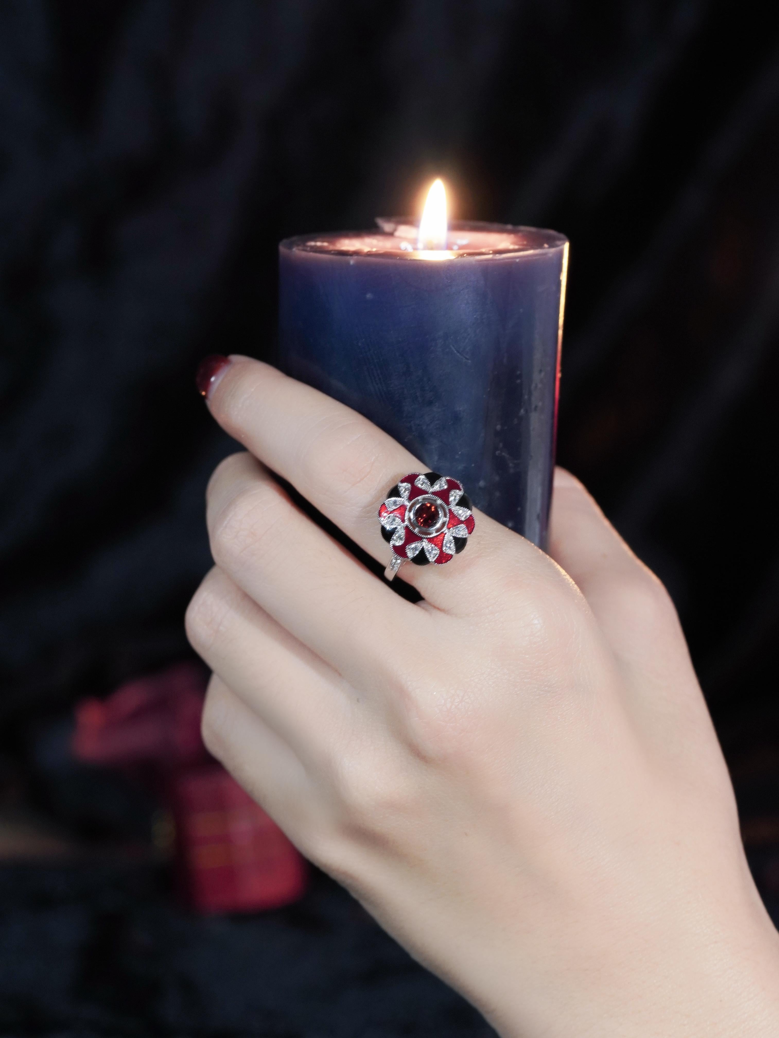 Ein Meisterwerk an Farbe und Handwerkskunst. Ein reicher runder Granat steht im Mittelpunkt, flankiert von zarten Rubinen im Dreiecksschliff, die eine feurige Note verleihen. Der Edelstein ist von einem Halo aus leuchtenden weißen Saphiren umgeben,