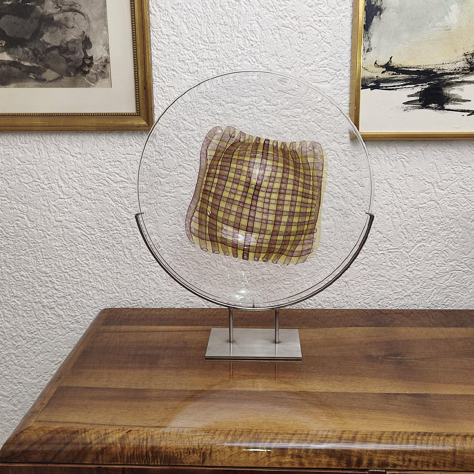 Gary Beecham, Großer dekorativer Glasteller „Textile-Gefäß“, 1982
Farbloses Kamee-Glas, mit zweischichtigem Bandmuster in der Mitte, wie ein gelb und braun kariertes Tuch. Halterung aus gebürstetem Edelstahl.
Signiert und datiert unter dem Boden