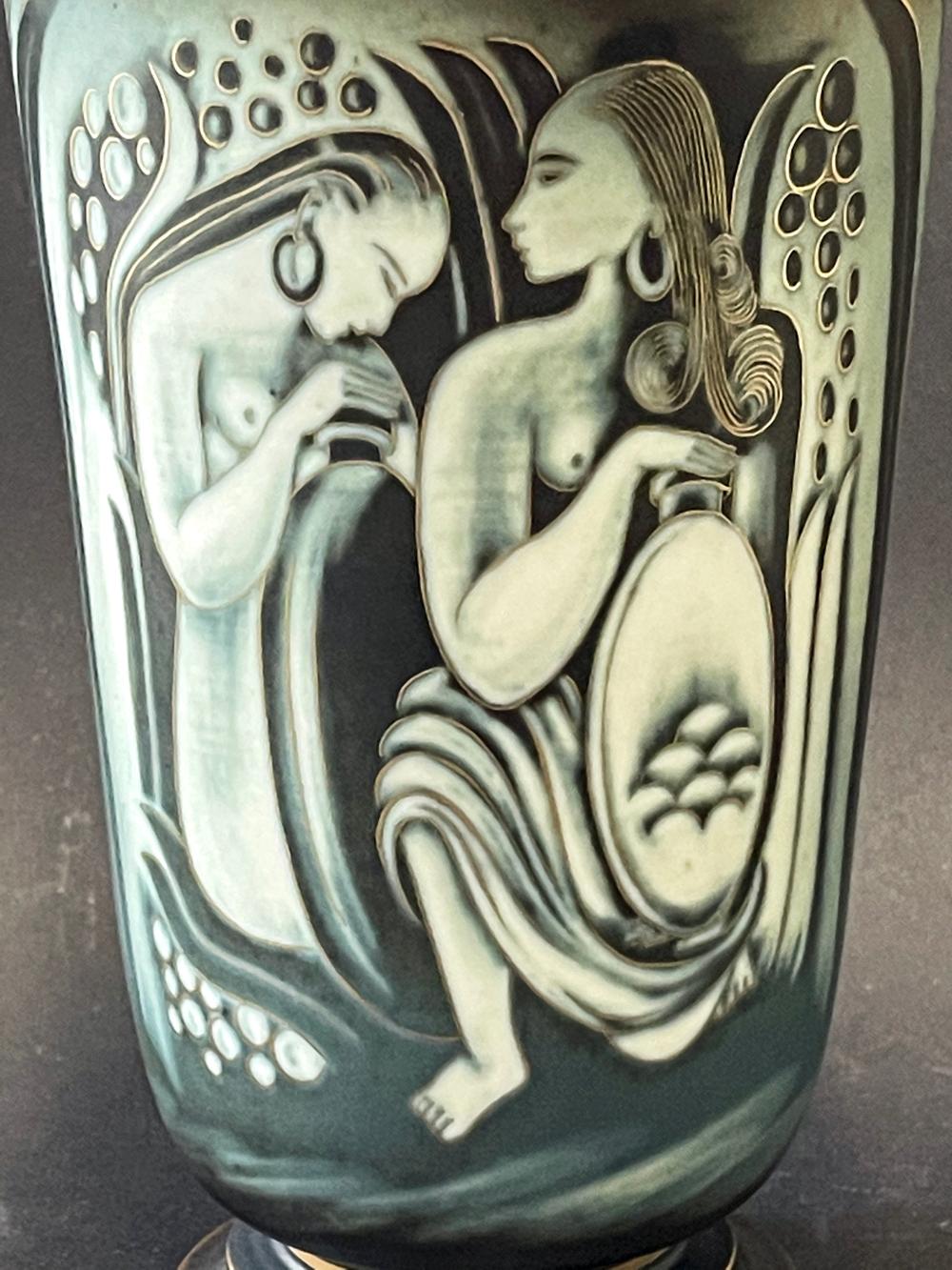 Diese elegante Vase, die von Gunnar Nylund für die berühmte schwedische Porzellanmanufaktur Rorstrand entworfen wurde, zeigt zwei nackte Frauenpaare, die am Rande eines von Schilf und üppigem Laub gesäumten Baches Wasser sammeln. Die Vase ist ein