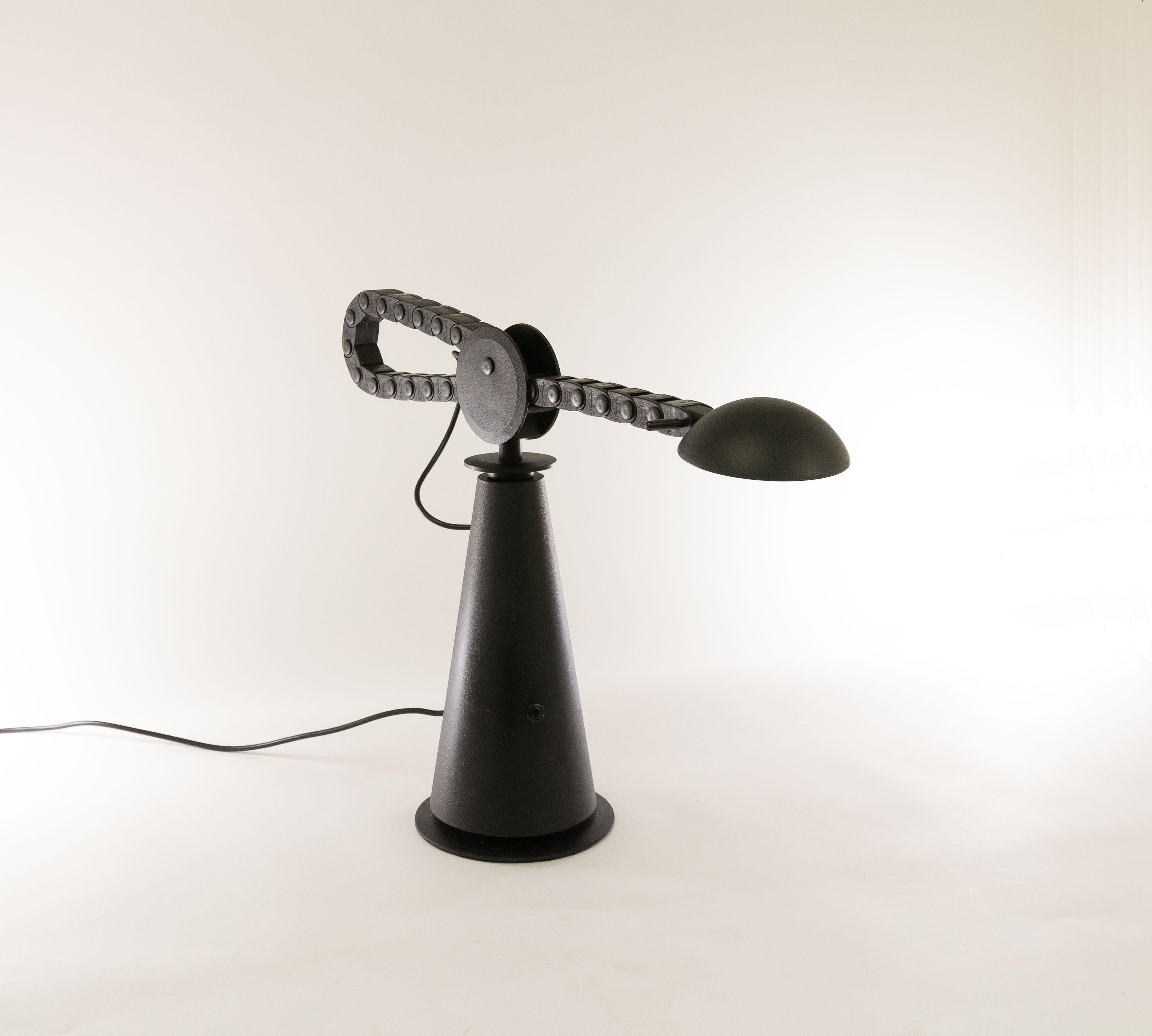 Gaucho halogen table lamp designed by Studio PER and manufactured by Egoluce in 1982.

The relatively heavy base is made of metal, just like the reflector and the 'wheel'. The adjustable arm which resembles a bicycle chain is made of