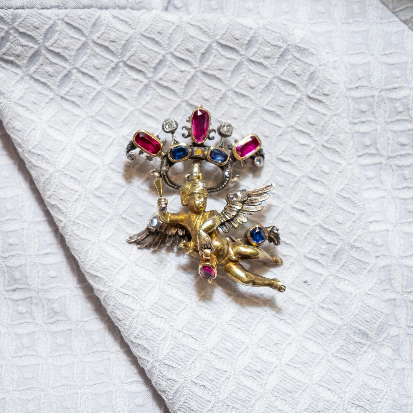 An antique cherub pendant, designed as a cherub in flight, holding an oval cut ruby, with articulated wings with diamond detail, with an oval cut sapphire set to a flowing piece of cloth, suspended from a ruby, sapphire and diamond crown, all