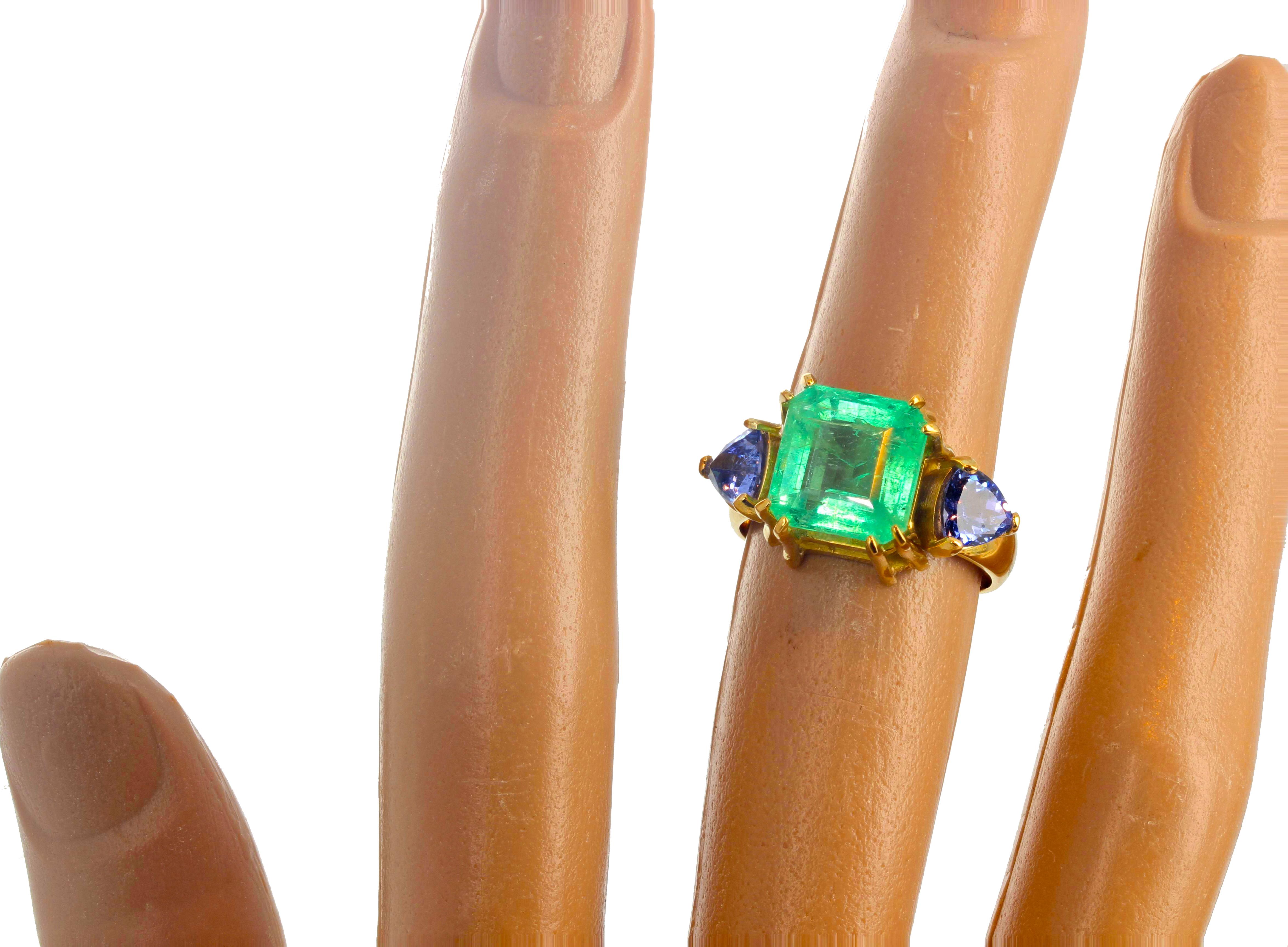 This spectacular natural center Columbian Emerald gemstone is 2.8 carats - 9.65mm x 9.30 mm -  enhanced with 6 mm x 6 mm trillion cut natural bright blue Tanzanites.  Handmade 18Kt yellow gold setting size 7 (sizable).  Please note that this