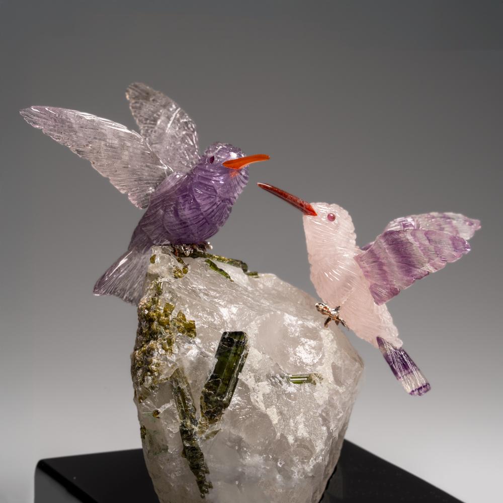 Cette sculpture de deux colibris est une pièce d'art unique, sculptée dans de la fluorine de qualité musée, avec des yeux en grenat et un bec en cornaline. Le colibri est perché au sommet d'une tourmaline verte dans une matrice de quartz et monté