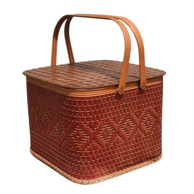 A red woven wicker picnic basket with metal handles, wood lid, and short tabletop for serving drinks or sandwiches. Created from woven red and brown wicker, the side of this basket is decorated in a diamond pattern around the body. The bottom