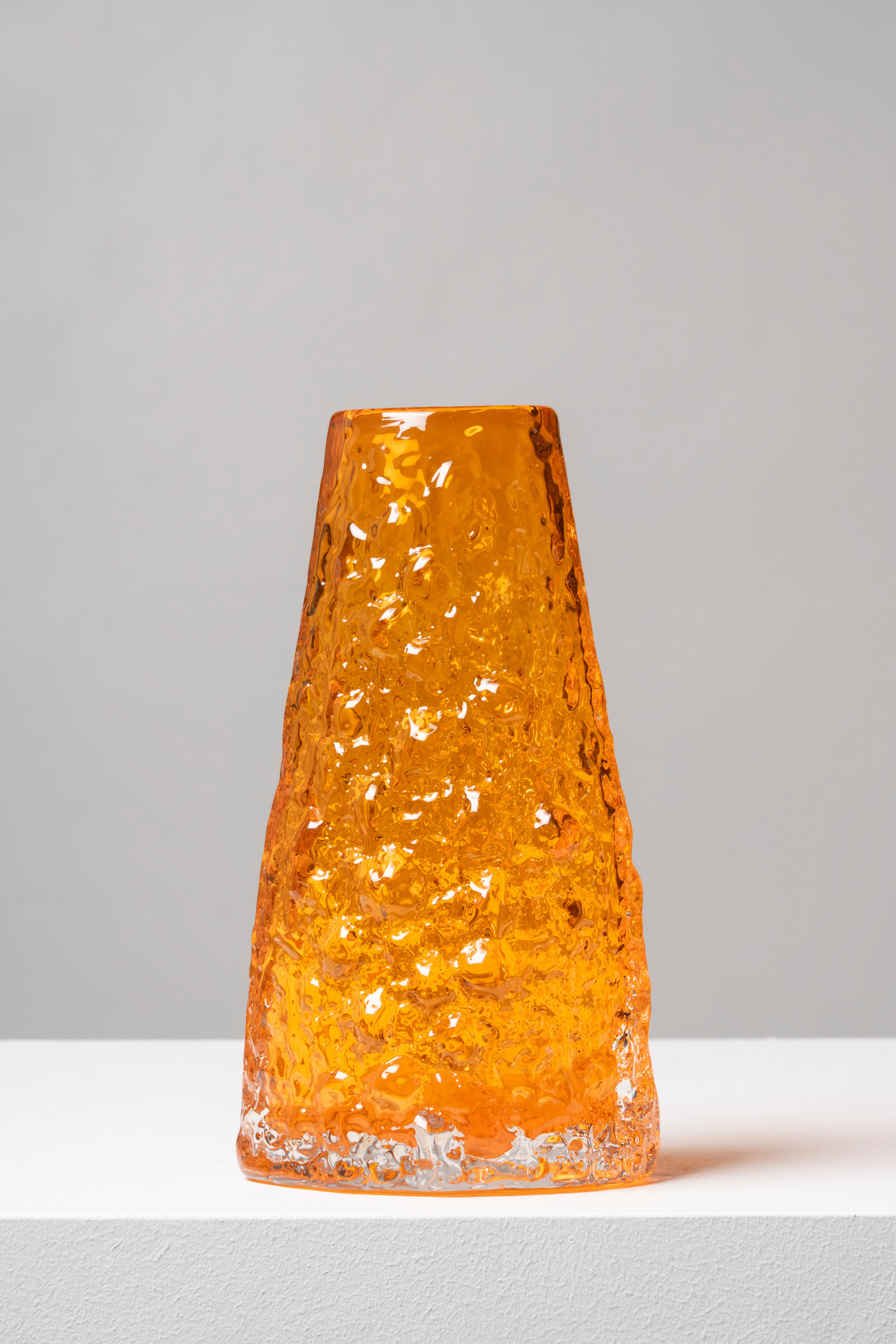 Orange textured glass vase with a flared base that narrows towards the mouth, following the organic shape of a volcano. The design of this piece was the brainchild of Geoffrey Baxter, who joined Whitefriars in 1954, bringing a renewed air to the