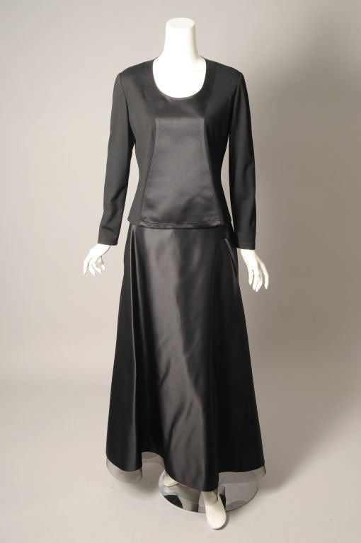 A striking two piece evening dress from Geoffrey Beene is done in a combination of black wool jersey and black satin. The center part of the top and the entire skirt are made from the satin. The rest of the top is jersey. The skirt is finished with