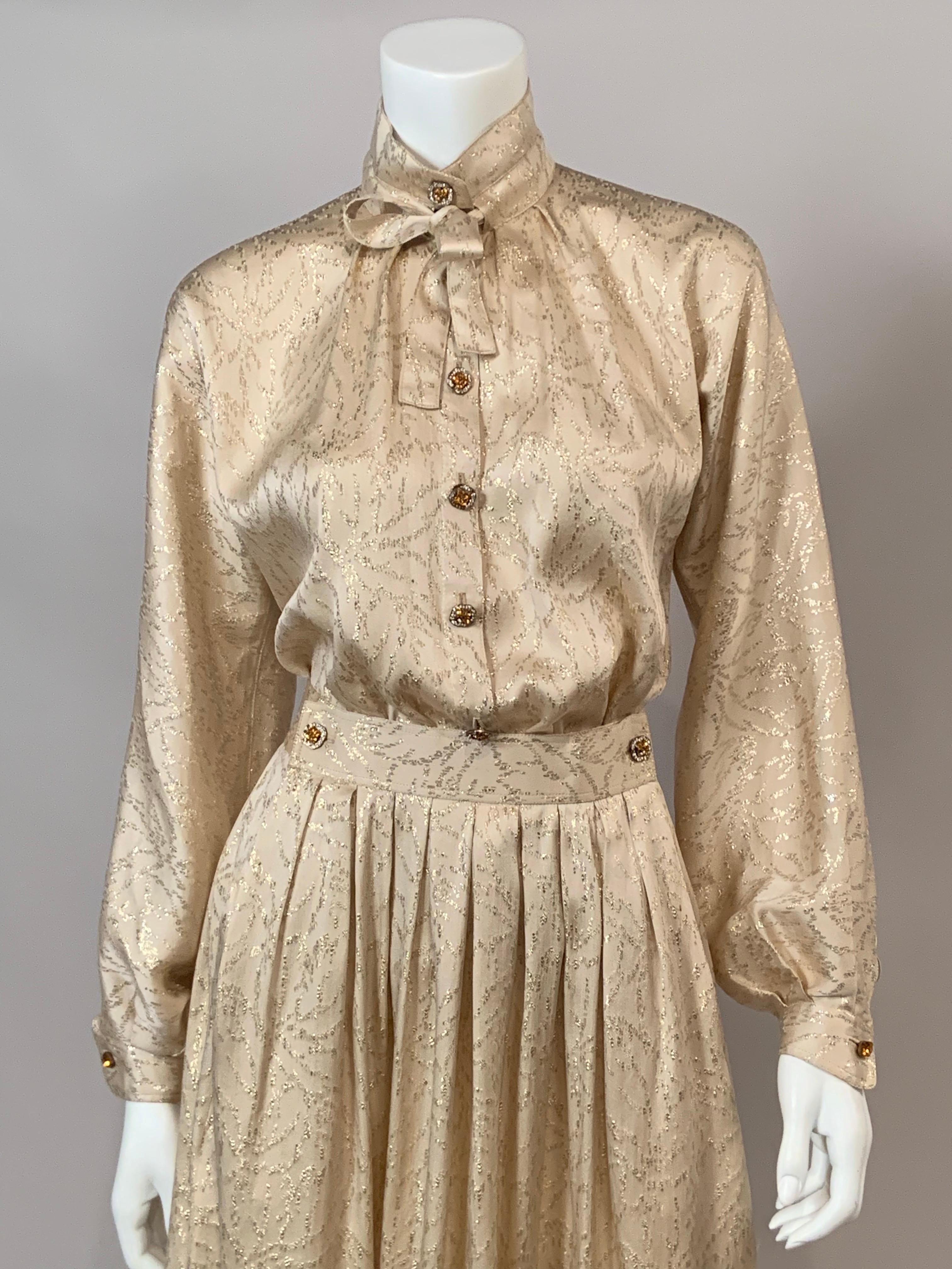 Geoffrey Beene a conçu cette robe deux pièces, chemisier et jupe, dans un style décontracté en utilisant un tissu et des boutons élégants.  Le tissu est de la soie crème avec une abondance de fils métalliques dorés dans un motif de feuilles. Le