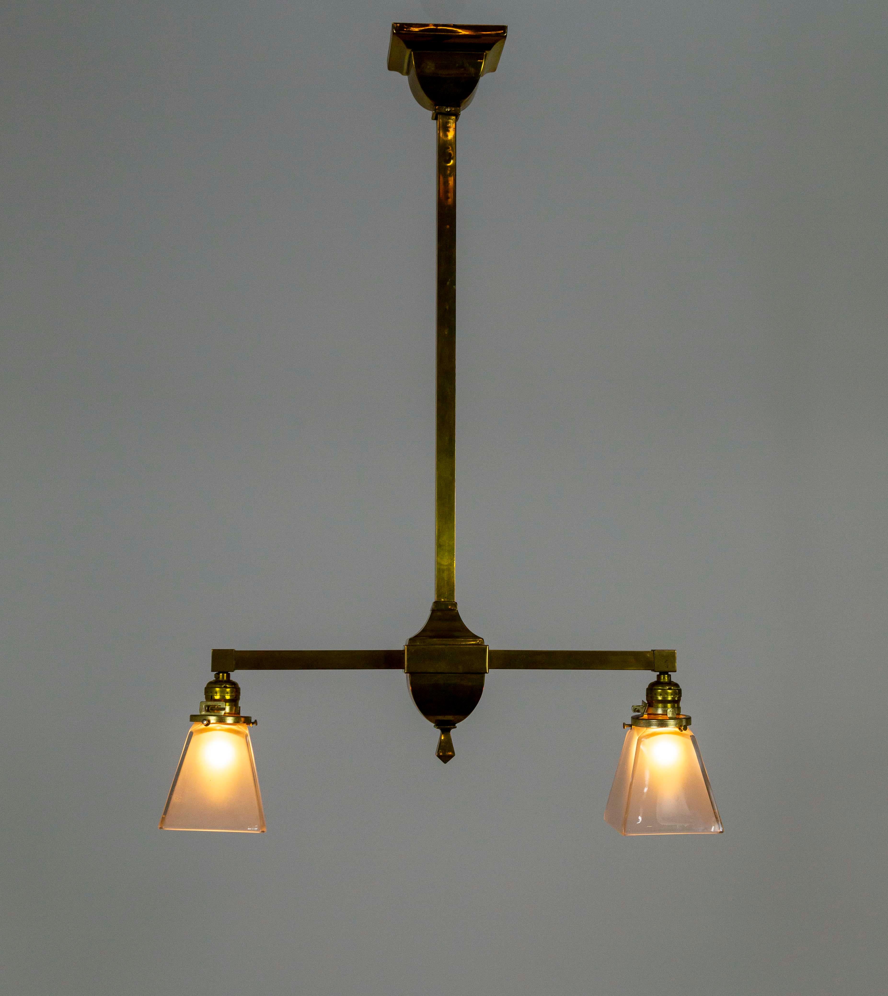 This simple, straight arm, brass chandelier is an excellent example of the Arts & Crafts movement. It has a streamlined design with squared stem and arms, and the brass itself has a lovely, patinated finish. The glass shades have a glossy exterior