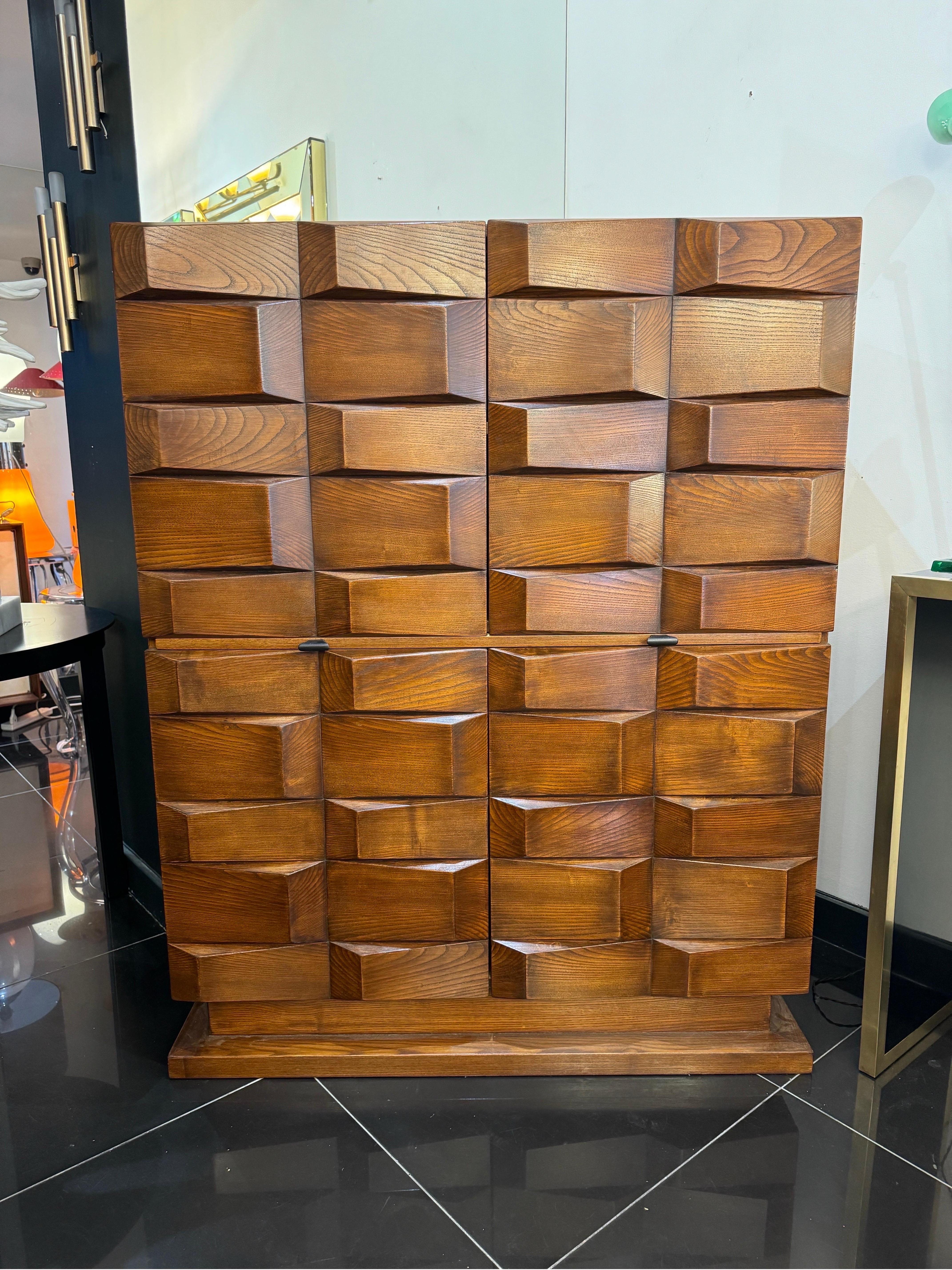 Un mobile bar in rovere scuro dal design geometrico e scolpito, con quattro ante a soffietto che rivelano un ampio spazio interno con un ripiano centrale estraibile nascosto. 

