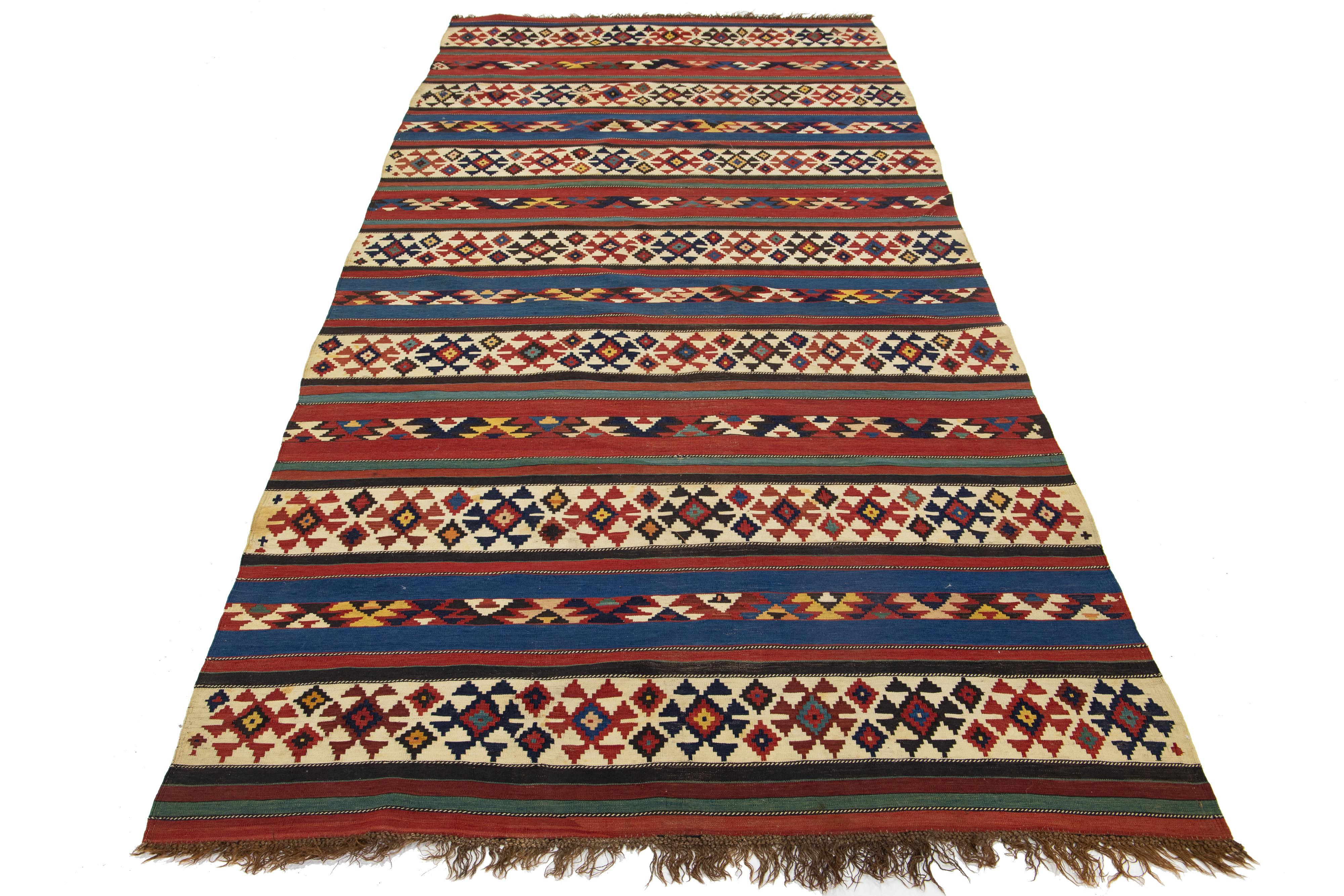 Presentamos una impresionante alfombra de lana kilim vintage, meticulosamente anudada a mano con un cautivador diseño geométrico sobre un llamativo fondo beige, azul y rojo.

Esta alfombra mide 4'11