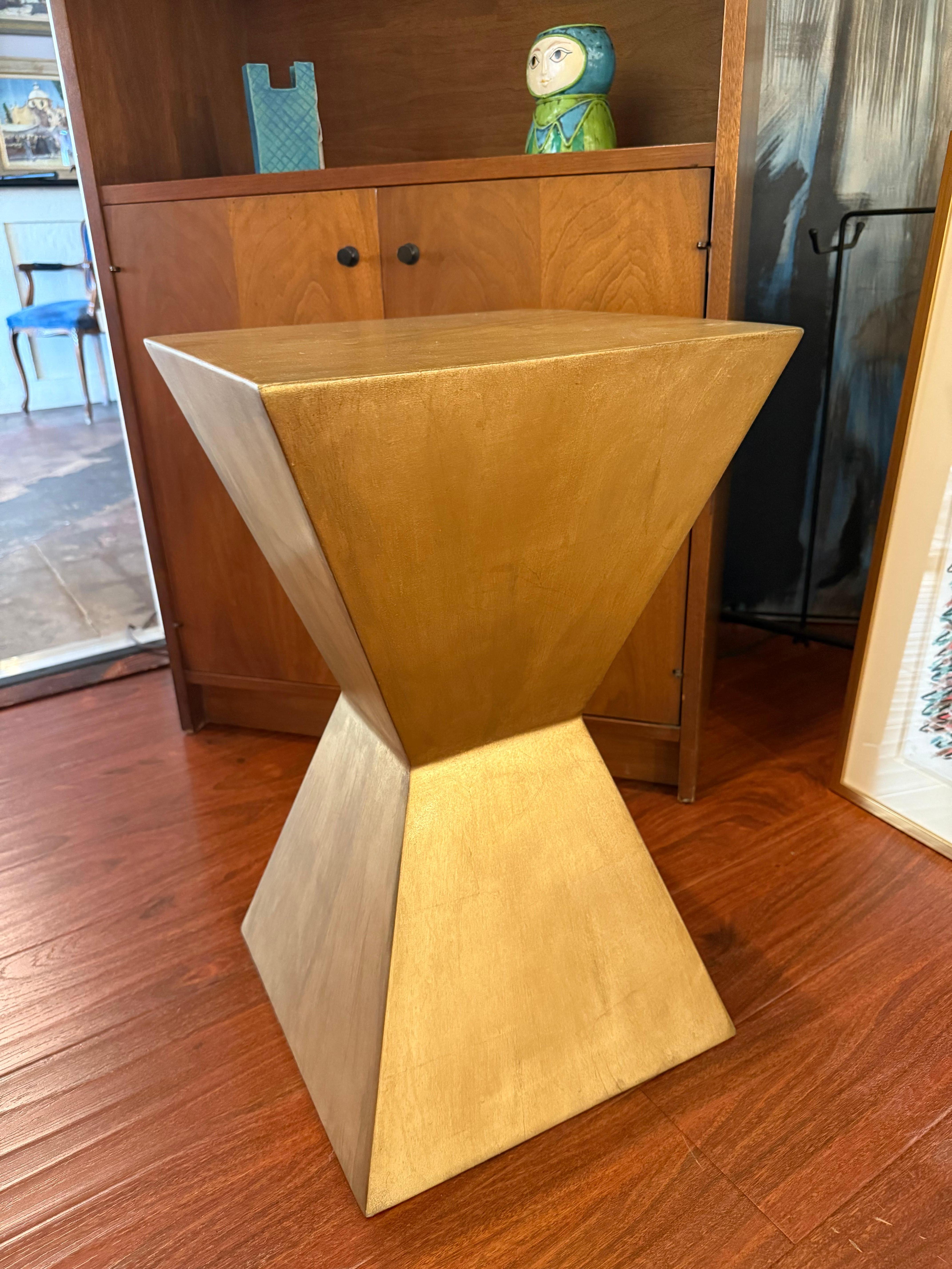 Questo piedistallo contemporaneo presenta un'audace silhouette a doppia piramide rifinita in un luminoso metallo color oro antico. I suoi piani angolari e la sua presenza scultorea incarnano il design di lusso moderno.