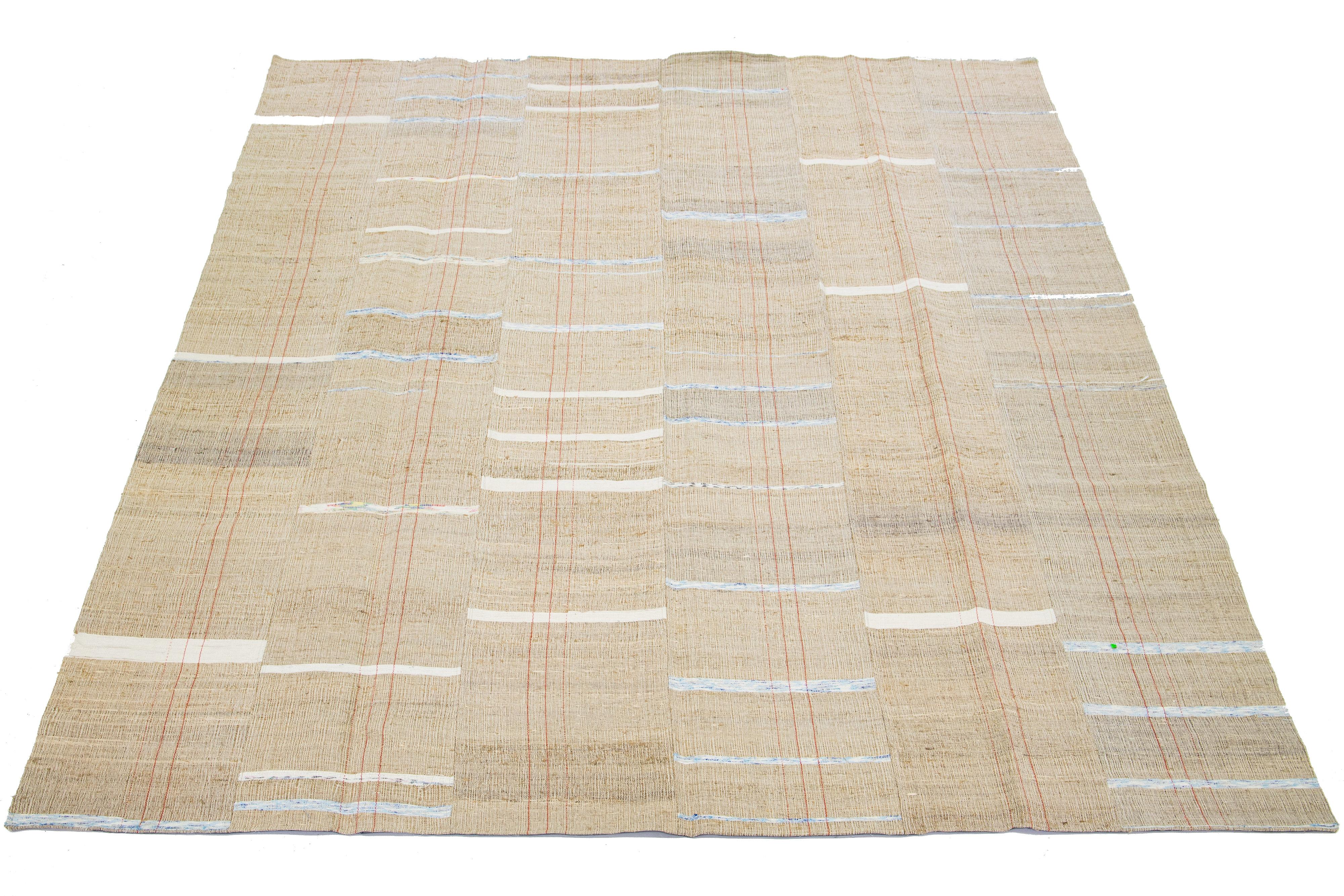 Dieser indische Teppich zeigt ein modernes Flachgewebe im Kilim-Stil, das aus Wolle gefertigt ist. Der Teppich hat ein beiges Feld mit einem gestreiften Muster in Weiß-, Blau- und Rosttönen.

Dieser Teppich misst 9'1