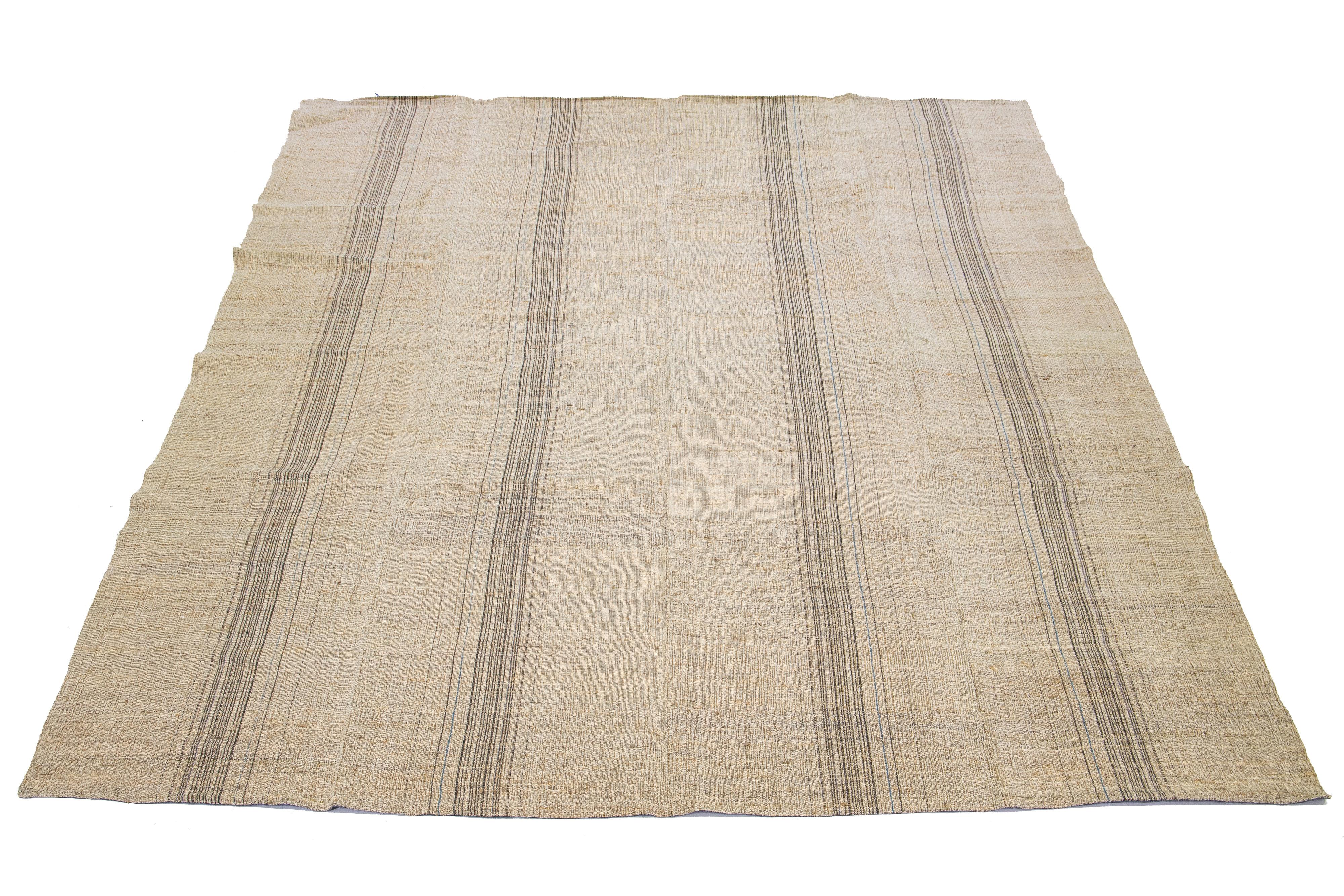 This Indian rug showcases a contemporary Kilim flatweave style crafted from wool. The rug has a light brown field with a striped pattern in blue and black shades.

This rug measures 10' x 13'3