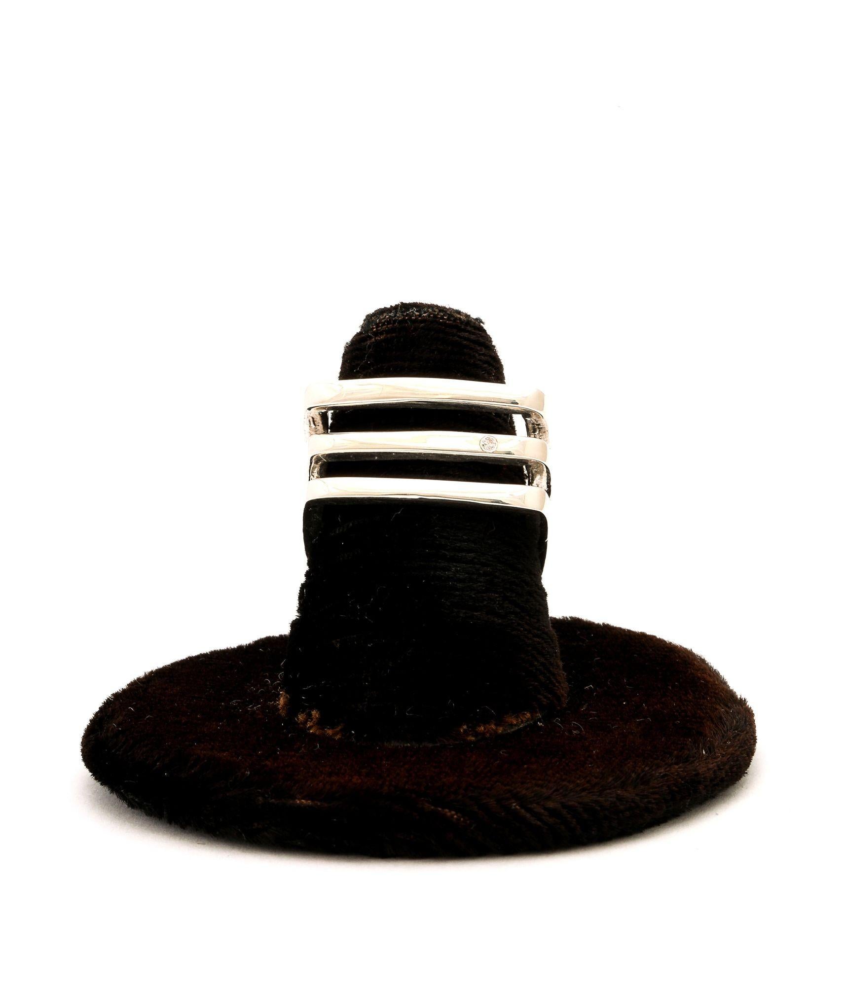 Un audaz anillo modernista de plata de ley maciza. Presenta una forma cuadrada y ancha con bandas paralelas y abiertas. Un pequeño diamante talla brillante está engarzado a ras para un brillo sutil.
Este anillo ofrece una fuerte estética geométrica