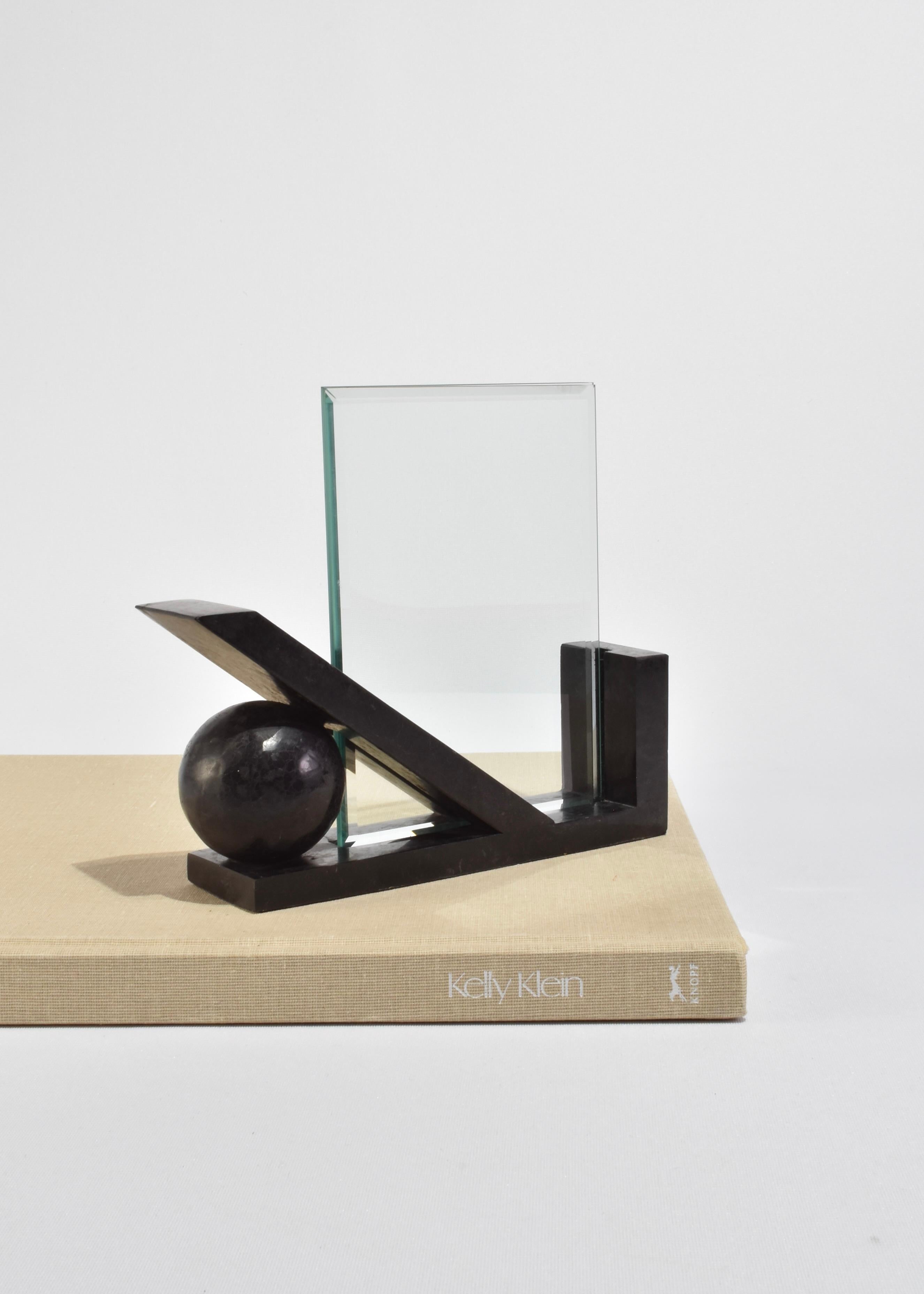 Un cadre photo sculptural composé de formes géométriques en pierre noire et verre transparent. La base angulaire supporte un panneau de verre rectangulaire, équilibré par une sphère solide et une base architecturale inclinée. À la fois fonctionnel