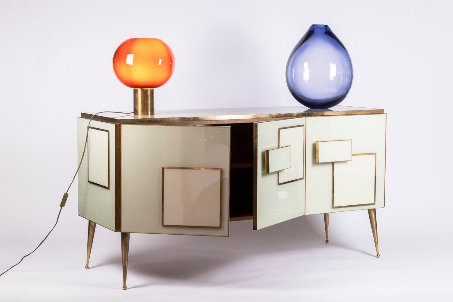 Sideboard in white colored glass and gilded brass, opening on the front with three doors and decorated with square and rectangular shapes, rectangular shapes used as handles. Spindle-shaped gilded brass base, enfing in small decorative