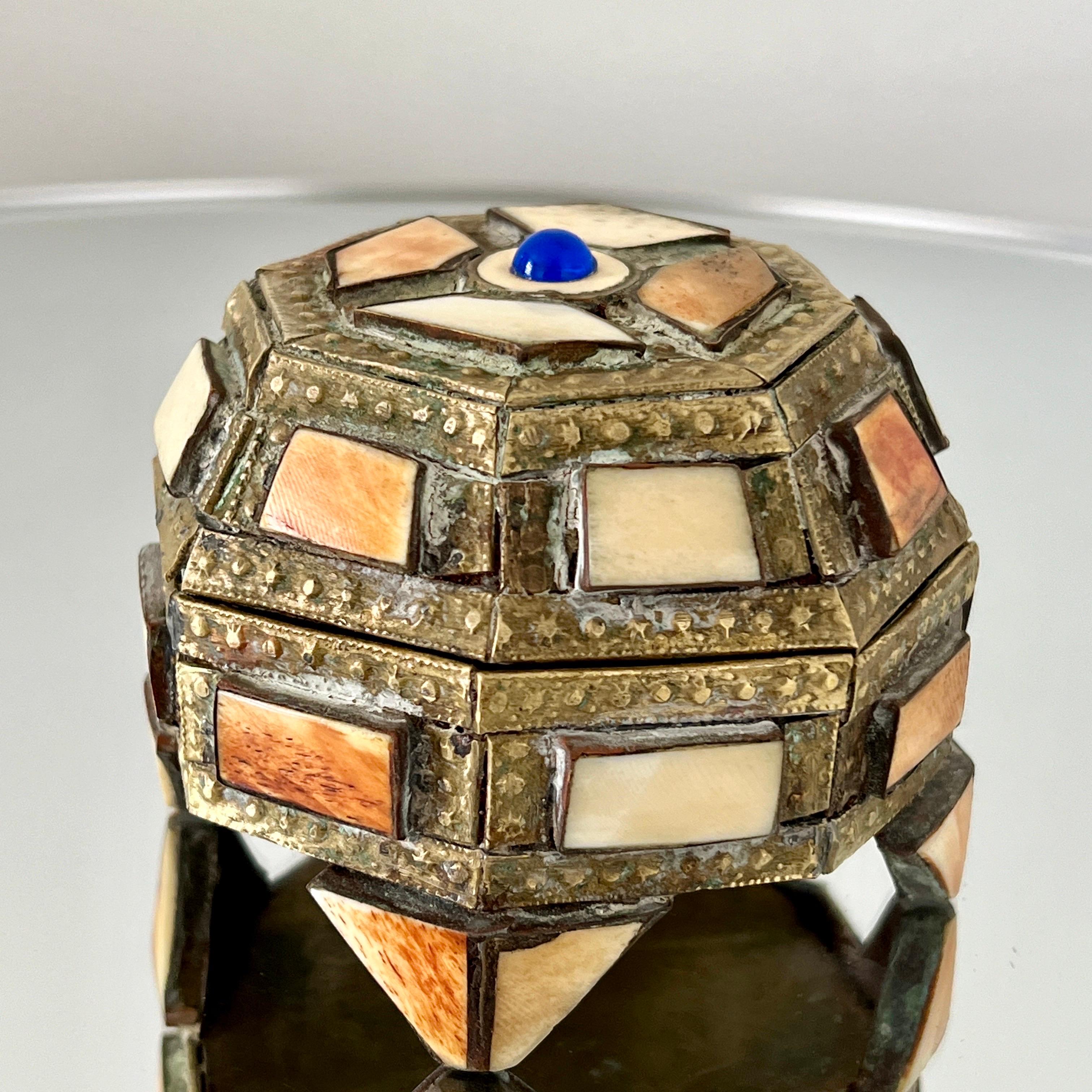 Handmade mosaic trinket box in beaded brass metal over wood with bone inlays.  The box has a geometrical octagon design with a footed base and lidded top.  It features incised metal motifs and a central semi-precious stone in blue.  The interior is