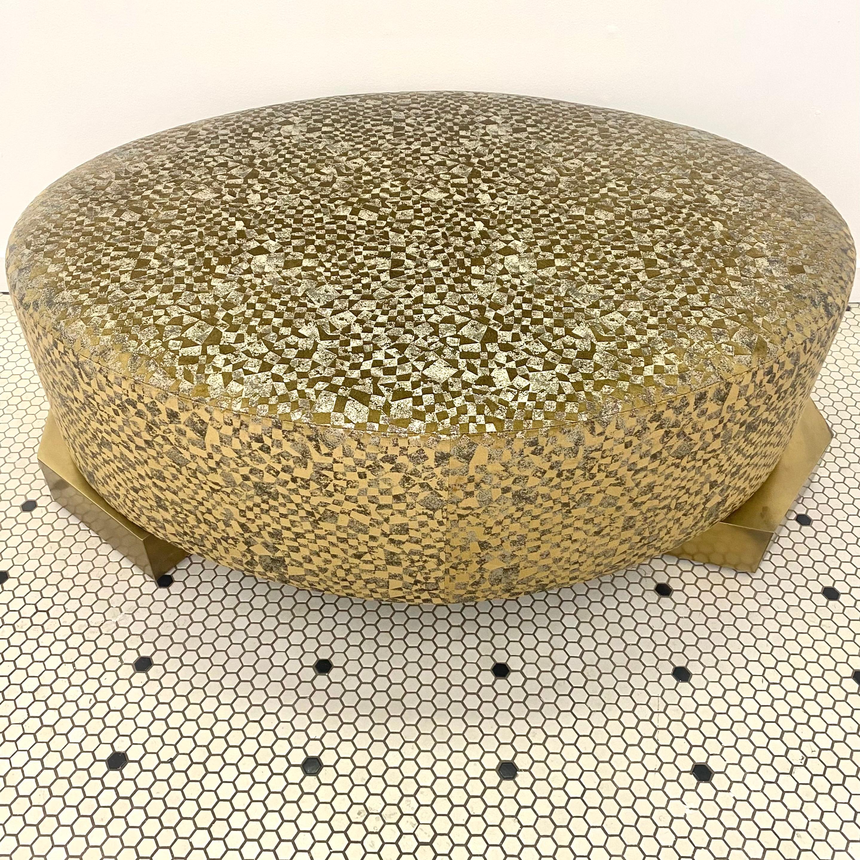 This stunning ottoman features a metallic mosaic fabric stretched to form a robust oval. The pouf cushion sturdily sits atop two asymmetrical, geometrical wood feet clad in brass. Light reflects off of the patterned fabric, and shifts in depth and