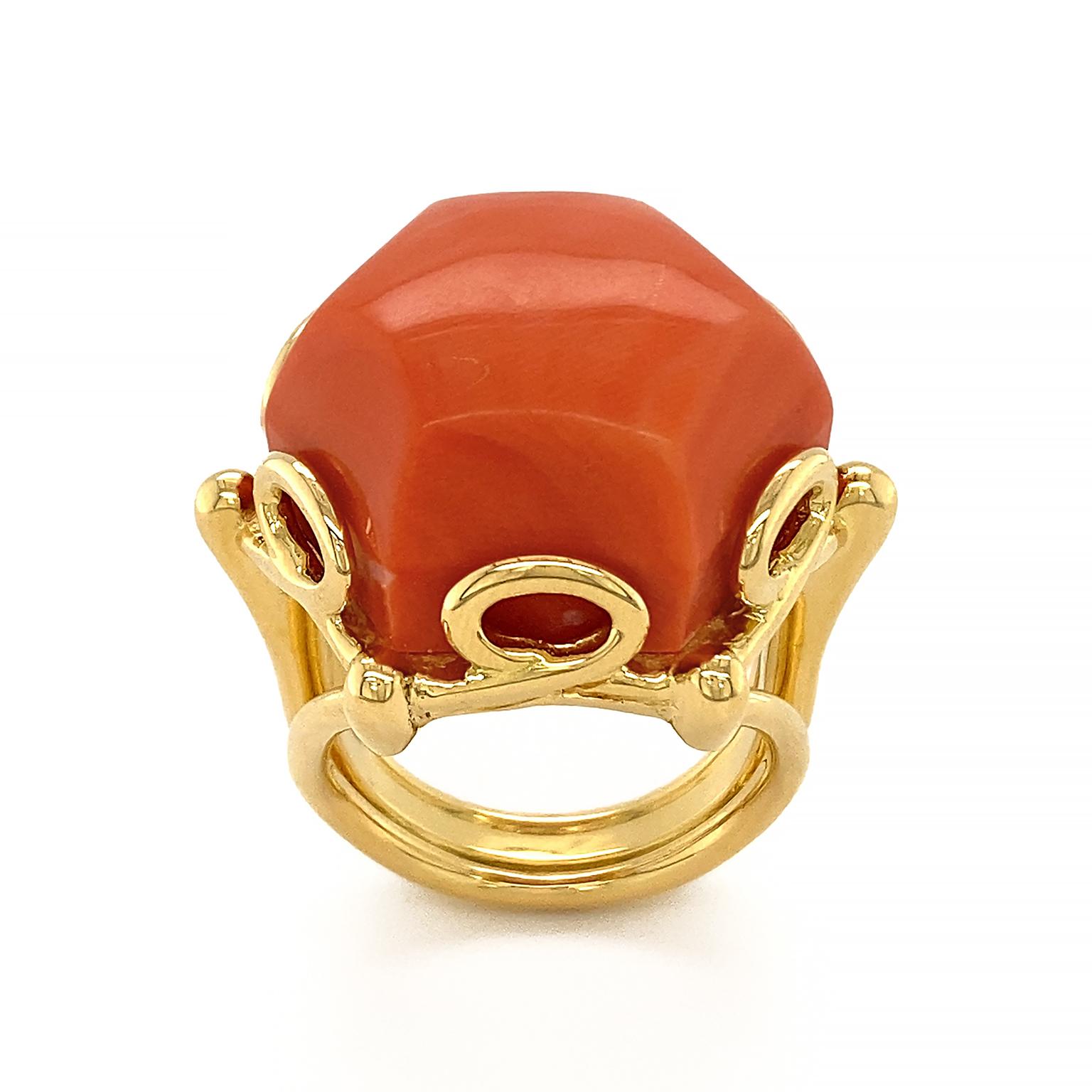 Geometrical Hexagon Dark Coral Solitaire Ring gleams with color. The deep orange-red jewel is carved into a hexagonal shape with tall sides. It rests on an 18k yellow gold band with a split shank. The setting of choice are gold loops which double as