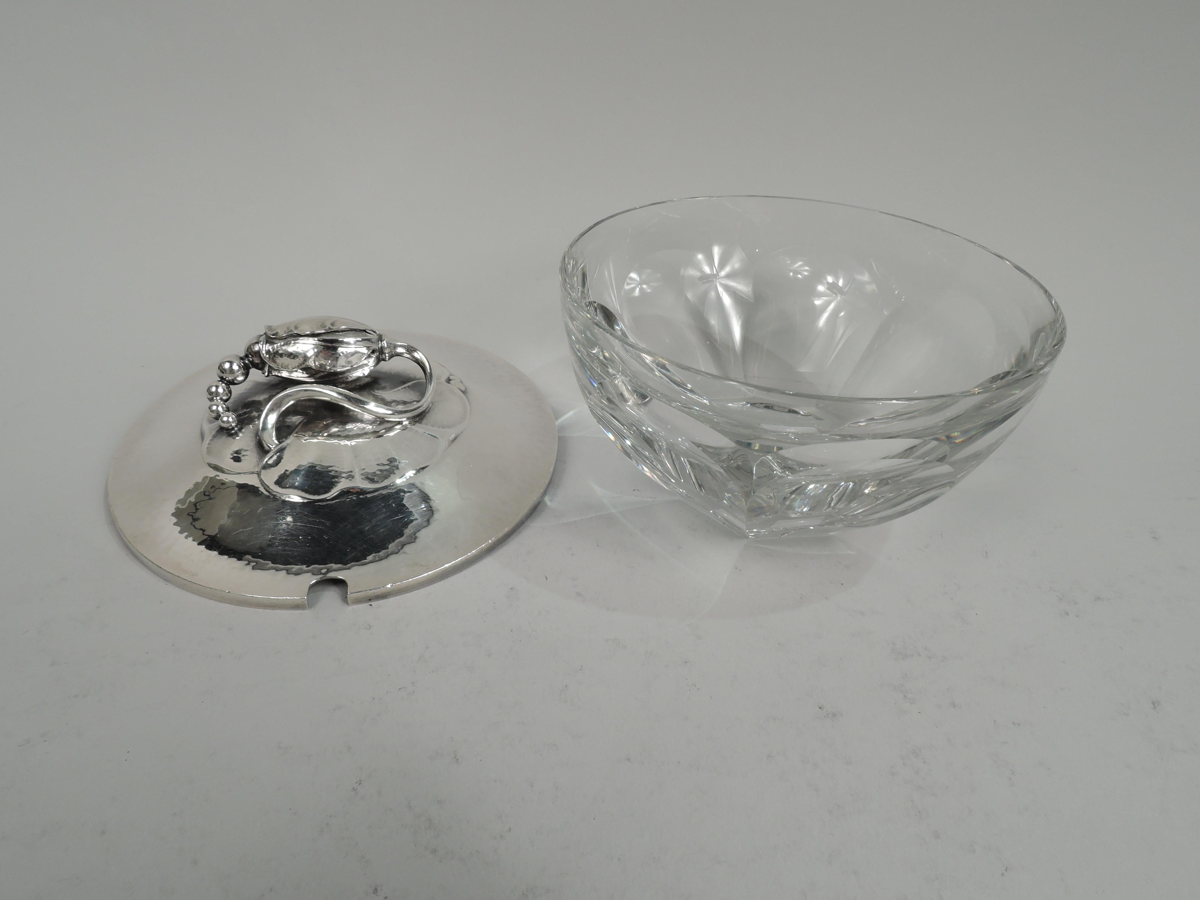 Blossom jelly bowl. Round crystal bowl with lobed and tapering sides. Cover sterling silver with gently raised and hand-hammered; on top is seed-spilling mounted to chased petal medallion. This design illustrated in Drucker, Georg Jensen, 1997, p.