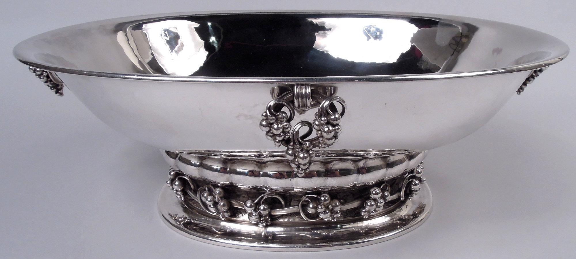 Dänischer moderner Tafelaufsatz aus Sterlingsilber. Hergestellt von Georg Jensen in Kopenhagen. Ovale Schale mit ausgestelltem Rand; an den Enden und Seiten sind lose Ringe mit Weintrauben angebracht. Der Sockel ist gelappt und auf einem gespreizten