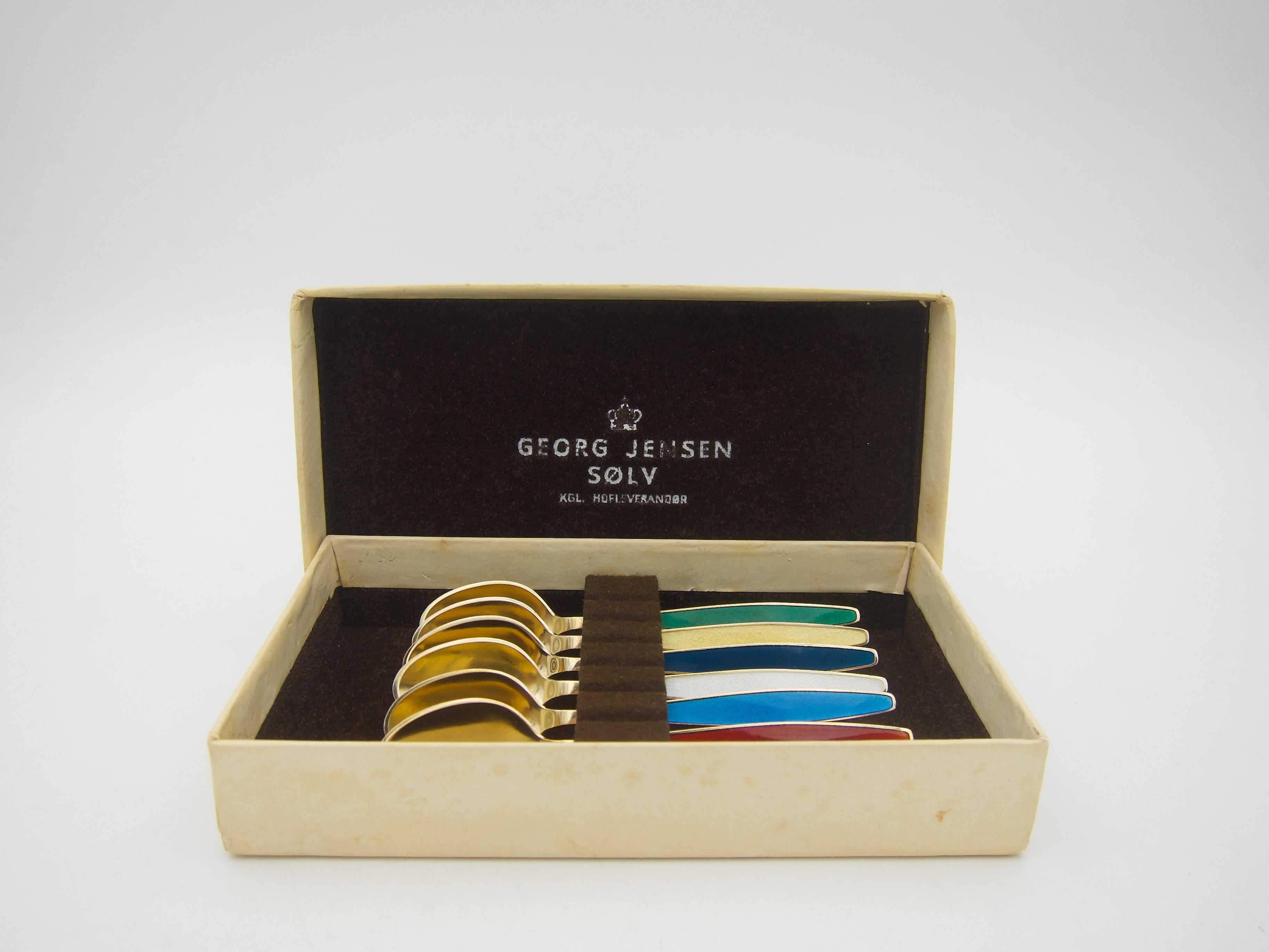 A vintage boxed set of six sterling silver gilt and enamel spoons from Georg Jensen, founded in Copenhagen, Denmark in 1904. Each solid silver spoon has a gold wash surface with a field of jewel toned enamel ornamenting the handles. The spoons date